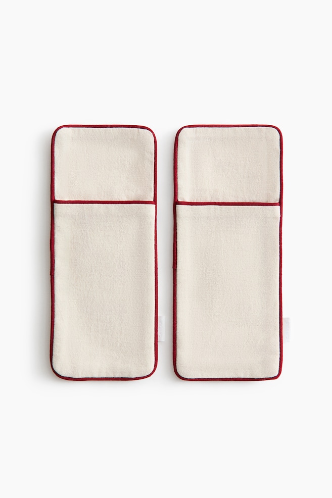 A pair of rectangular, cream-colored fabric cutlery holders is facing front side-by-side, showcasing a textured woven material. The holders are featuring front pockets and contrasting red piped borders along their rounded edges and pocket openings. - 1