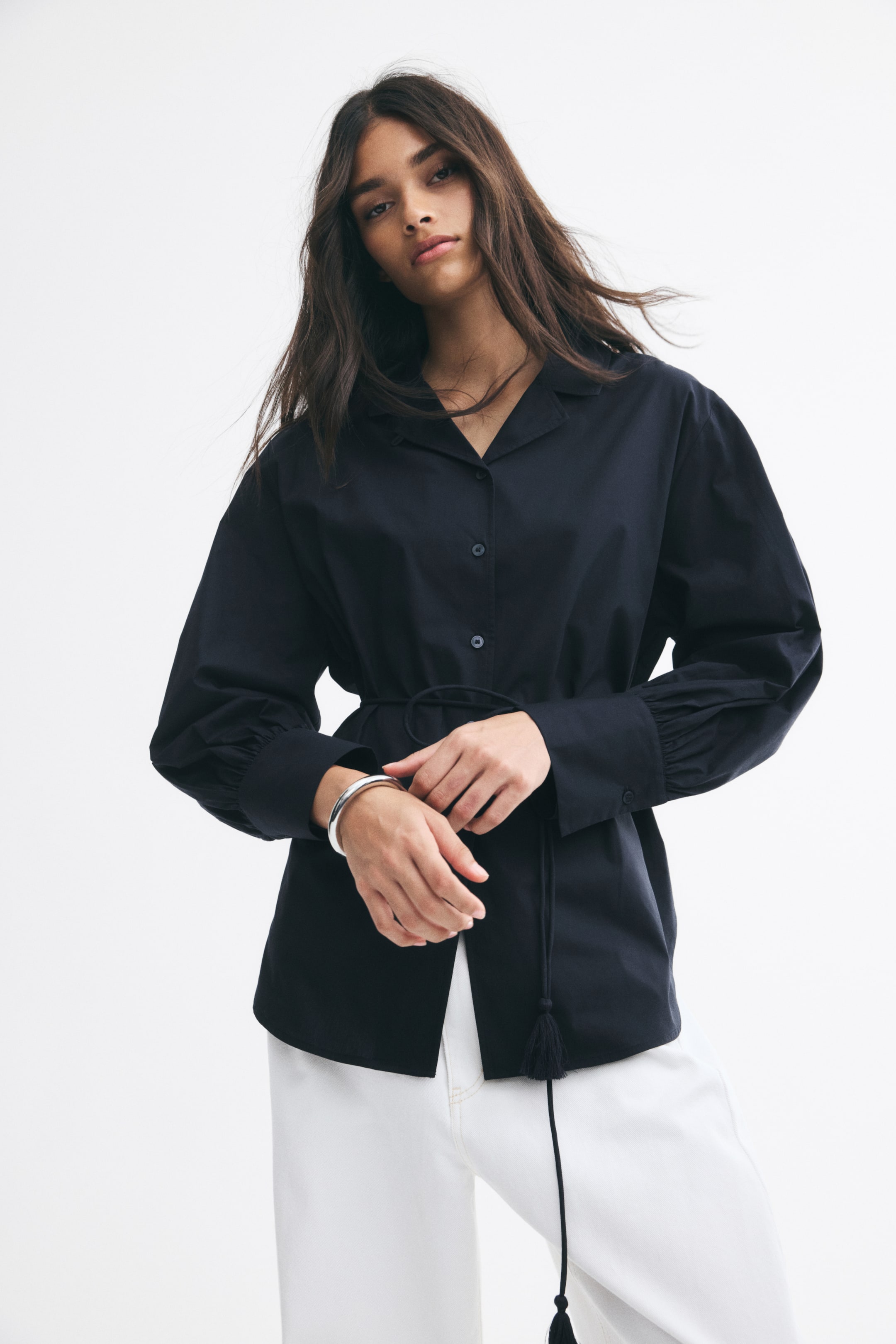 View larger image: A long black shirt in a relaxed fit, featuring a resort collar, button-down front, and dropped shoulders with voluminous sleeves, is worn by the model. The shirt is cinched at the waist with a slim tasselled tie belt and styled with white trousers.