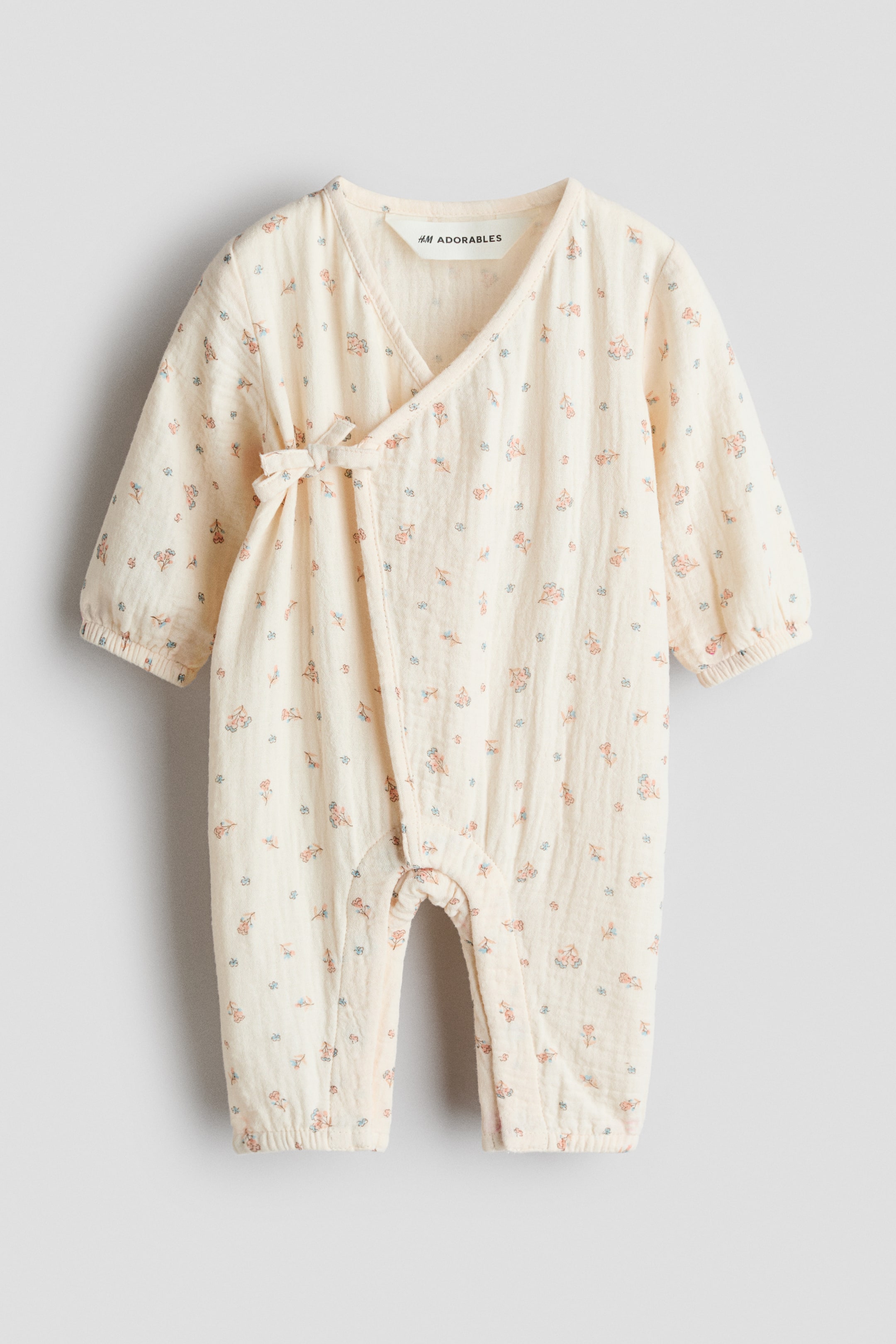 View larger image: This long-sleeved romper suit is displayed from the front, crafted from soft, light-coloured muslin fabric adorned with an all-over floral pattern. It features a wrapover front with narrow ties forming a bow on one side, while covered elastication gathers the cuffs and leg hems.