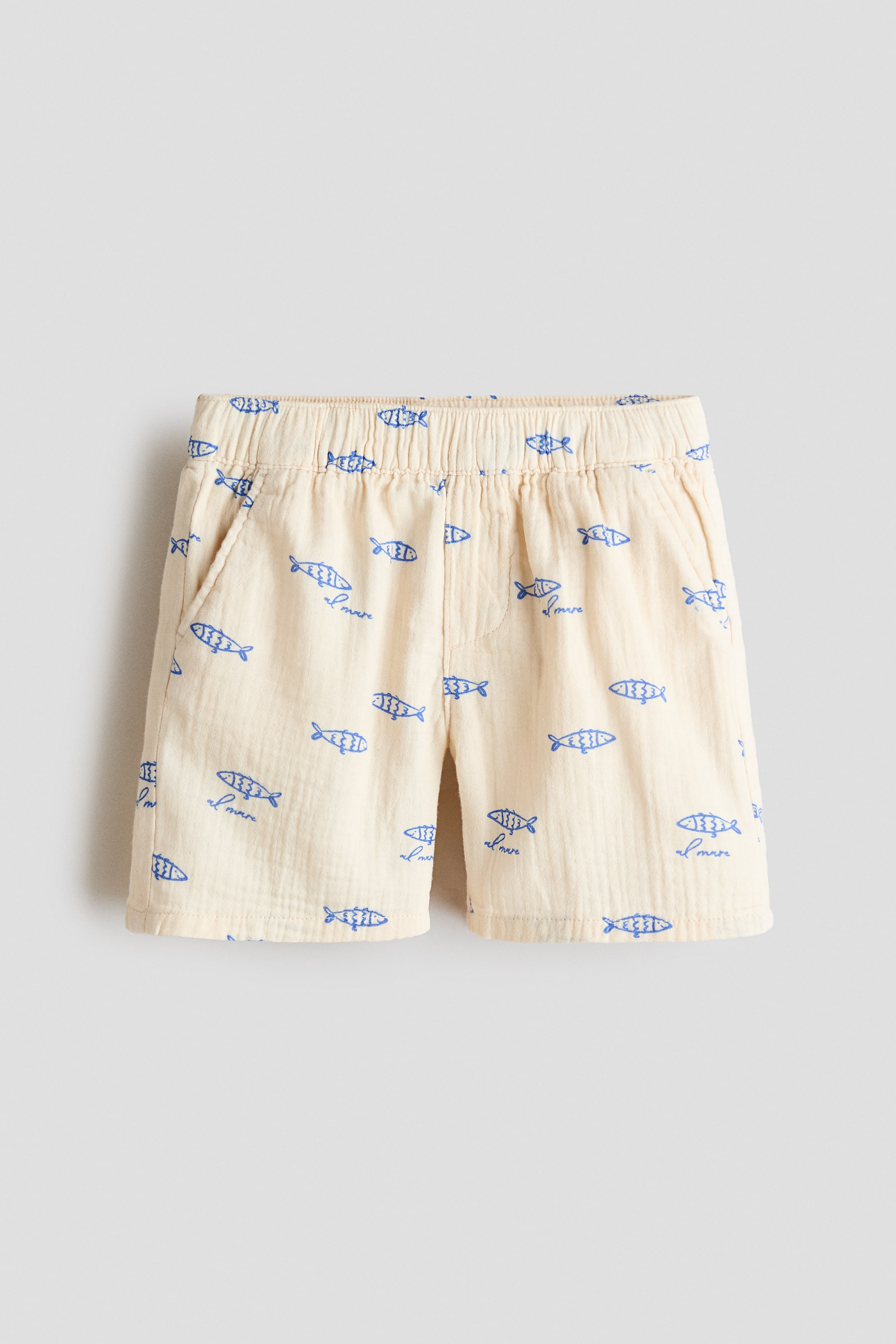 Größeres Bild ansehen: Knielange Shorts aus weichem, bedrucktem Baumwoll-Musselin mit einem durchgehenden blauen Fisch- und 'al mare'-Muster, das von vorne präsentiert wird. Sie verfügen über einen elastischen Bund, einen Fake-Schlitz und diagonale Seitentaschen.