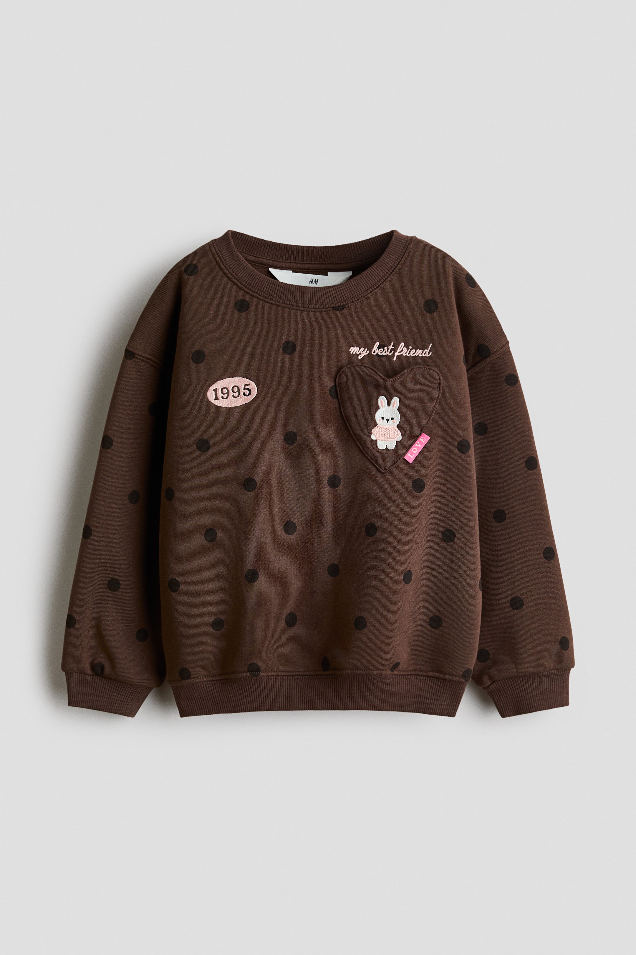 View larger image: A brown, relaxed-fit sweatshirt is shown from the front, featuring an all-over dark polka-dot pattern, dropped shoulders, and ribbed trimming at the neckline, cuffs, and hem. The chest is adorned with a pink oval patch displaying the year "1995" on one side, and a heart-shaped patch showcasing an embroidered bunny below the cursive text "my best friend" on the other.