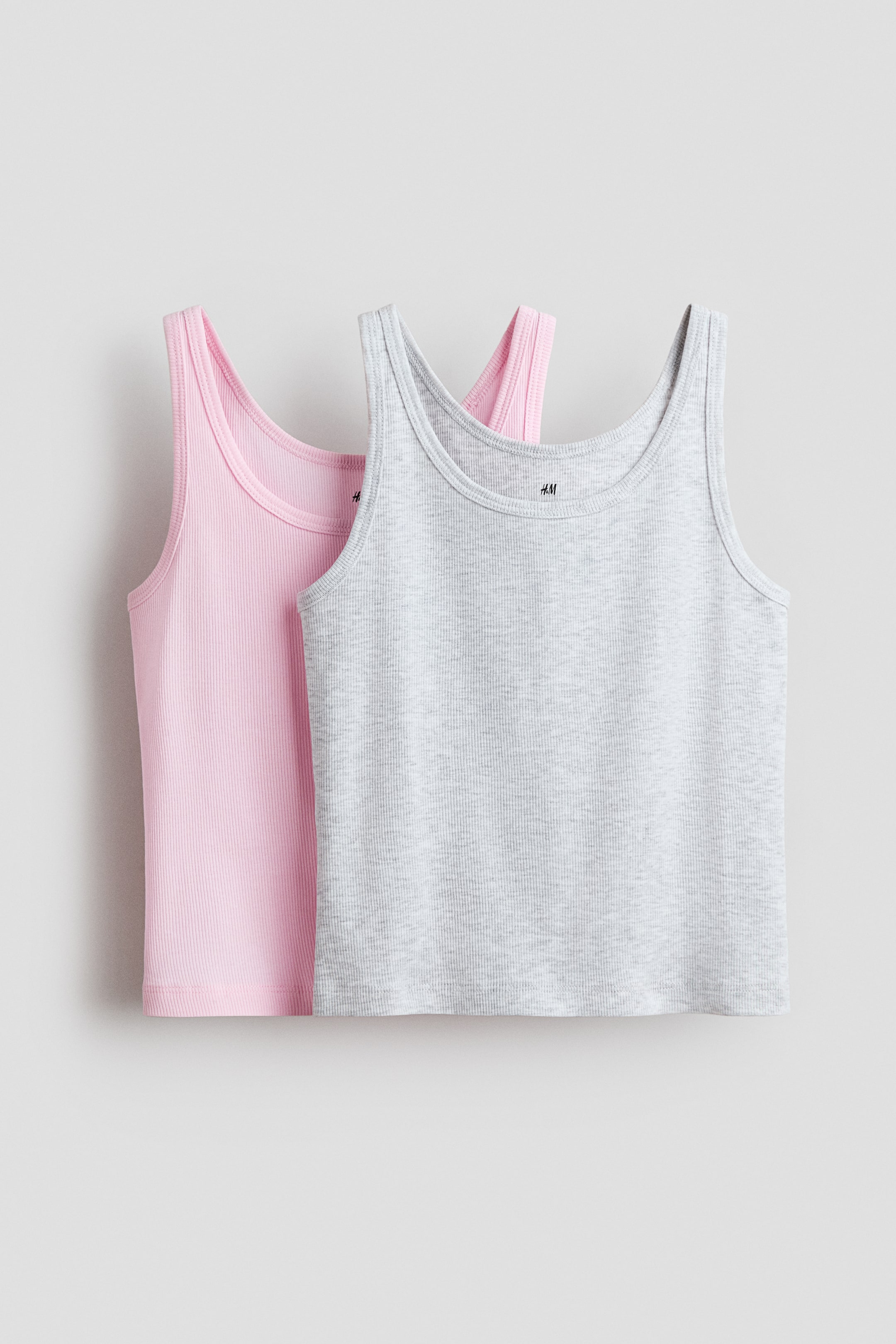 View larger image: Two tank tops are displayed: one in light gray and the other in light pink. Both feature a ribbed knit texture, scoop neckline, and wide shoulder straps, with the gray top slightly layered over the pink one.