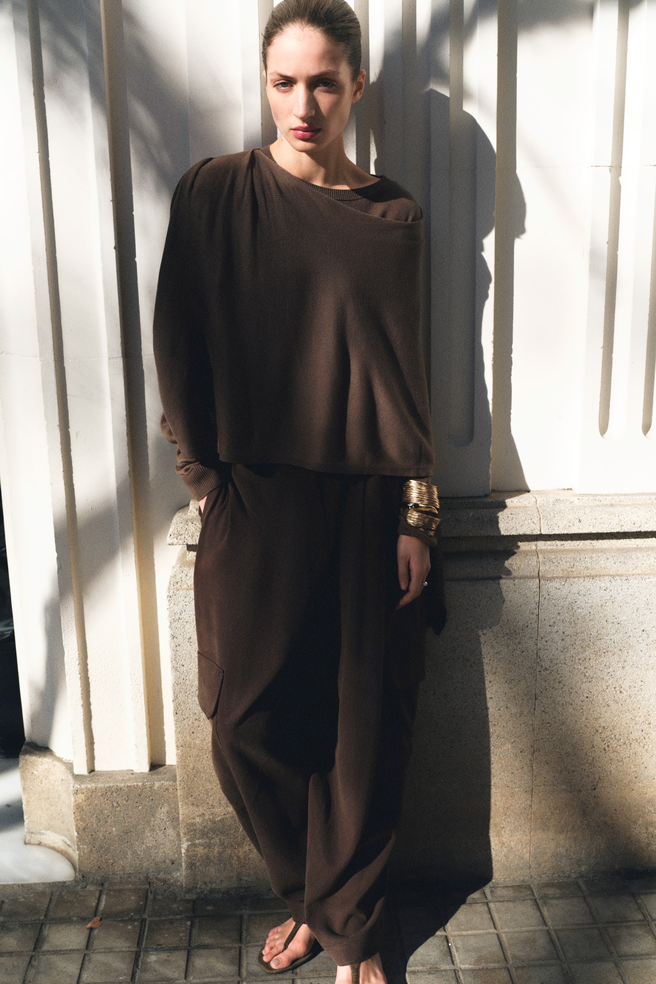 Grotere afbeelding bekijken: Een donkerbruin oversized gebreid topje met een off-shoulder halslijn wordt gedragen door het model, gecombineerd met bijpassende loszittende broek met een zijzak waar haar hand op rust. Gouden armbanden zijn gestapeld op haar pols, en bruine teenslippers sieren haar voeten.