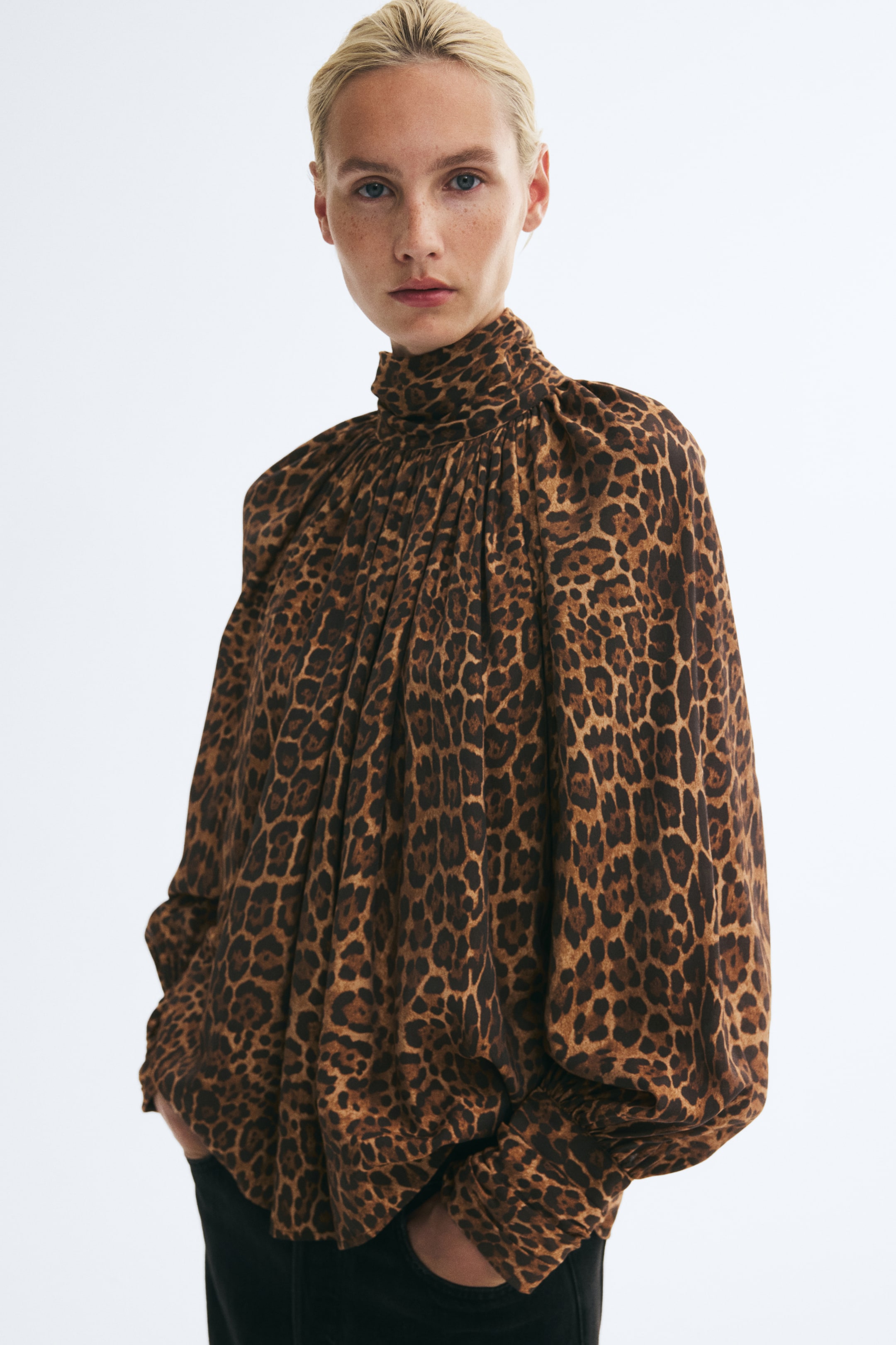 Prikaži veću sliku: Model nosi oversized bluzu sa leopard dezenom, sa naboranim uspravnim ovratnikom i naborima duž izreza, kao i voluminoznim balon rukavima sa širokim manžetnama. Bluza je stilizovana uz tamne pantalone, u koje model odmara ruke.