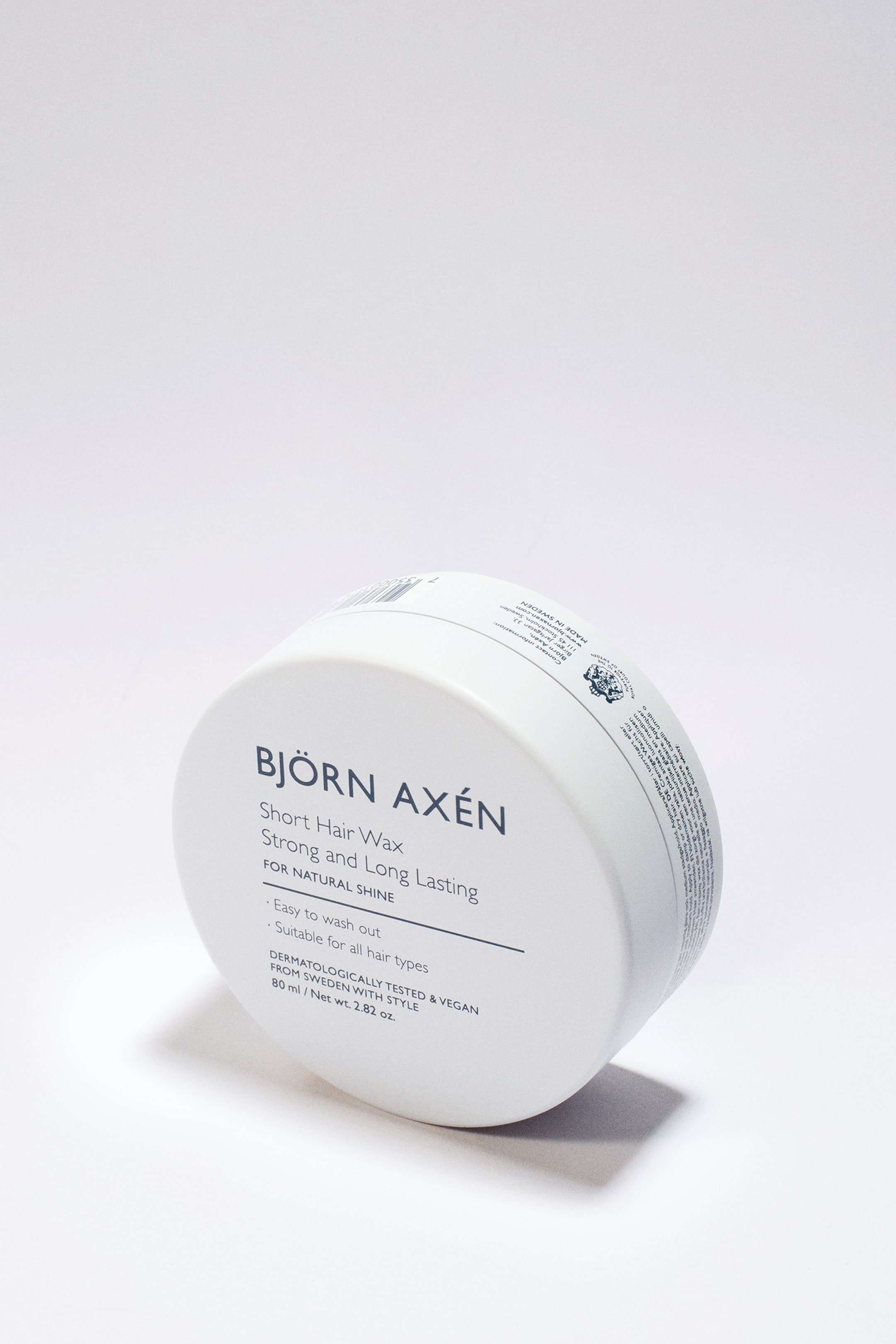 Se større billede: En hvid, rund hårvoksbeholder er vinklet fremad og til højre, hvilket fremhæver dens frontetiket med "BJÖRN AXÉN Short Hair Wax Strong and Long Lasting FOR NATURAL SHINE." Etiketten angiver også funktioner som "Let at vaske ud," "Velegnet til alle hårtyper," "DERMATOLOGISK TESTET & VEGANSK," og "80 ml / Nettovægt 2.82 oz."