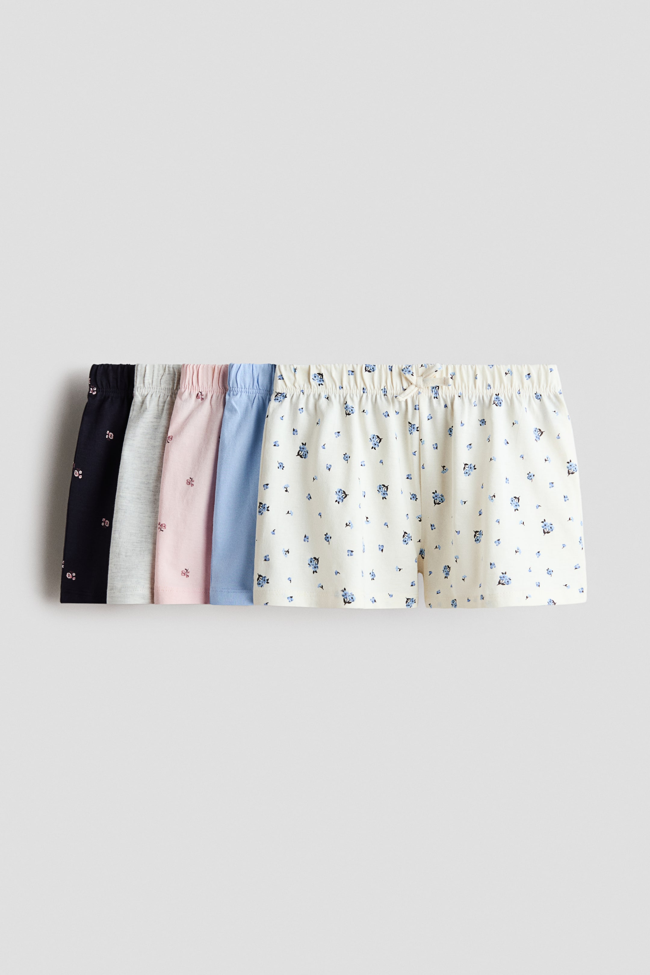 Visualizza immagine più grande: Cinque paia di pantaloncini sono esposti frontalmente, mettendo in risalto le loro cinture elastiche e i vari design. Questi includono un paio color crema con stampa floreale blu e un fiocco decorativo, un paio tinta unita azzurro, un paio rosa chiaro con una stampa discreta, un paio grigio melange e un paio blu navy con una stampa discreta.