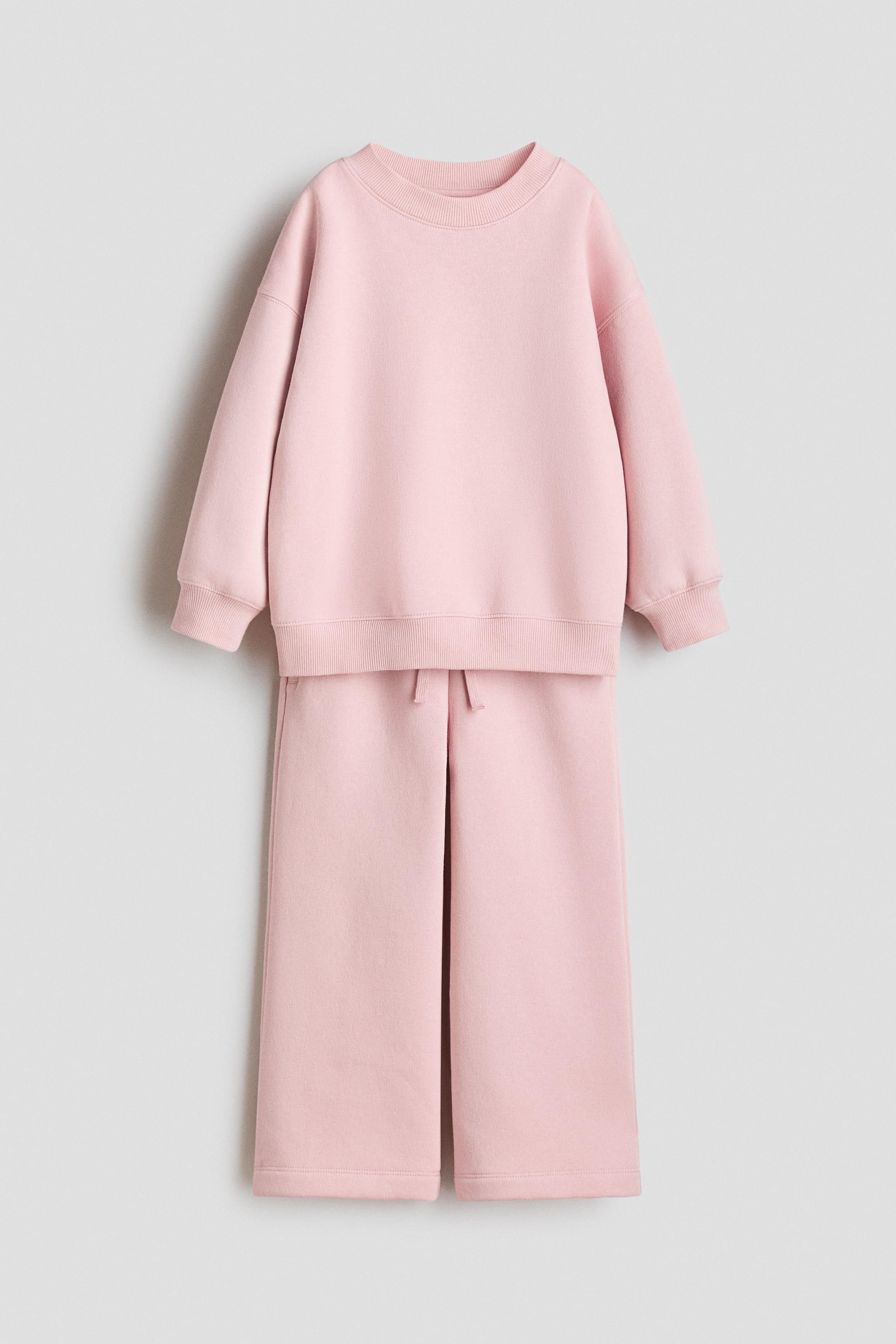 View larger image: A pink sweatshirt set is displaying its front, featuring a top with a round, ribbed neckline, dropped shoulders, long sleeves, and ribbed cuffs and hem, and coordinating joggers presenting an elasticated, drawstring waist, diagonal side pockets, and flared legs.
