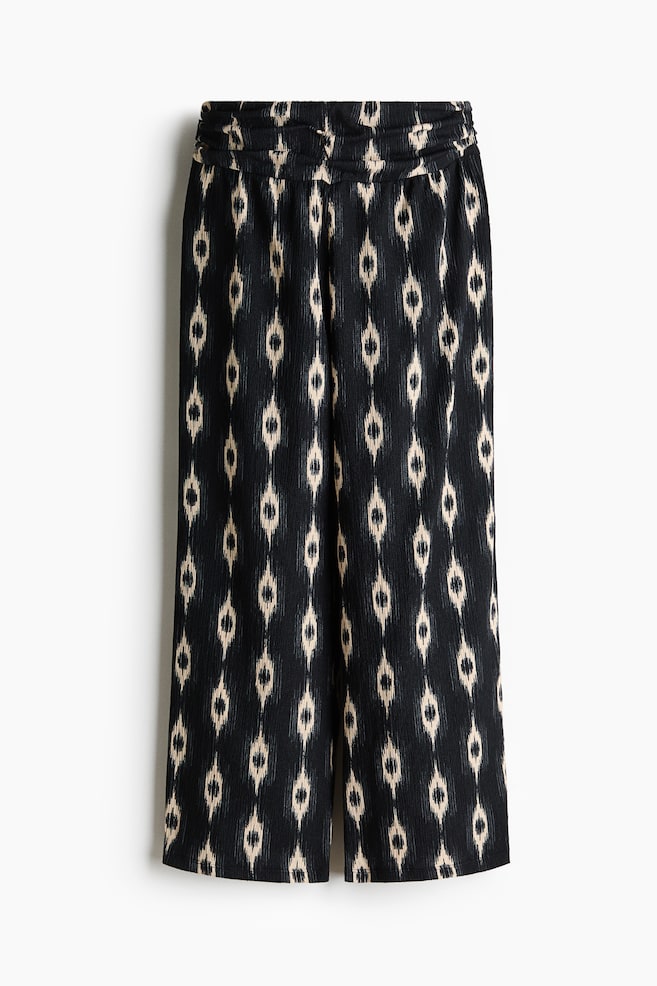 Los pantalones presentan un ajuste holgado y de pierna ancha en tejido de jersey arrugado, con una cintura alta y un cinturón fruncido, y un patrón de diamantes ikat en negro y beige claro, mostrado desde el frente. - 1
