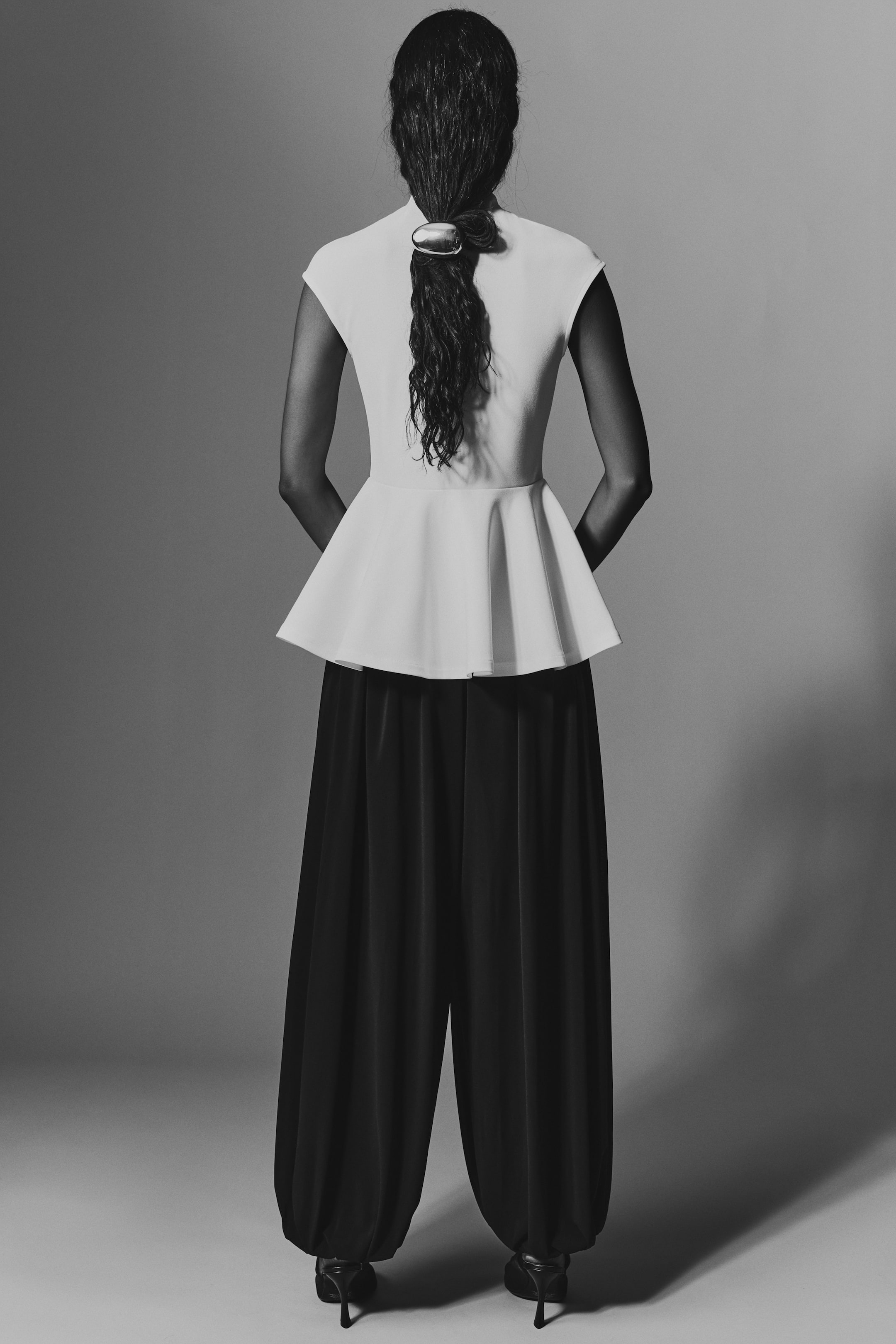 Ampliar la imagen: Blusa peplum blanca con mangas tipo capa y cuello alto, con un corpiño ajustado que se abre en un amplio volante peplum. Se muestra de espaldas, combinada con pantalones negros de pierna ancha que se fruncen a la altura de los tobillos sobre unos zapatos de tacón alto negros.