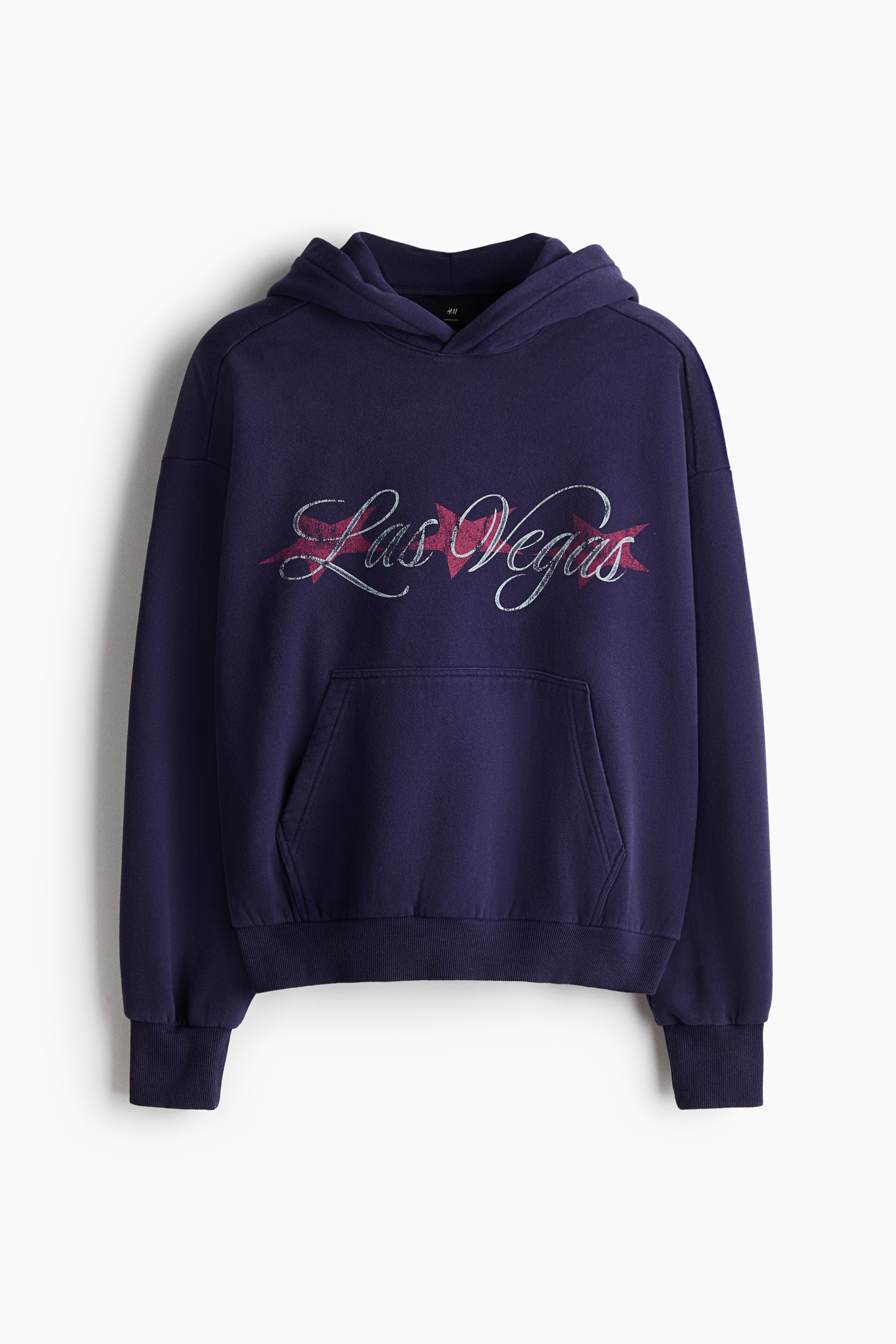 An oversized, dark purple hoodie featuring a graphic print of âLas Vegasâ in cursive script across the chest is being worn by the model. The hoodie features dropped shoulders, a kangaroo pocket, and ribbed cuffs, and is paired with black drawstring pants.