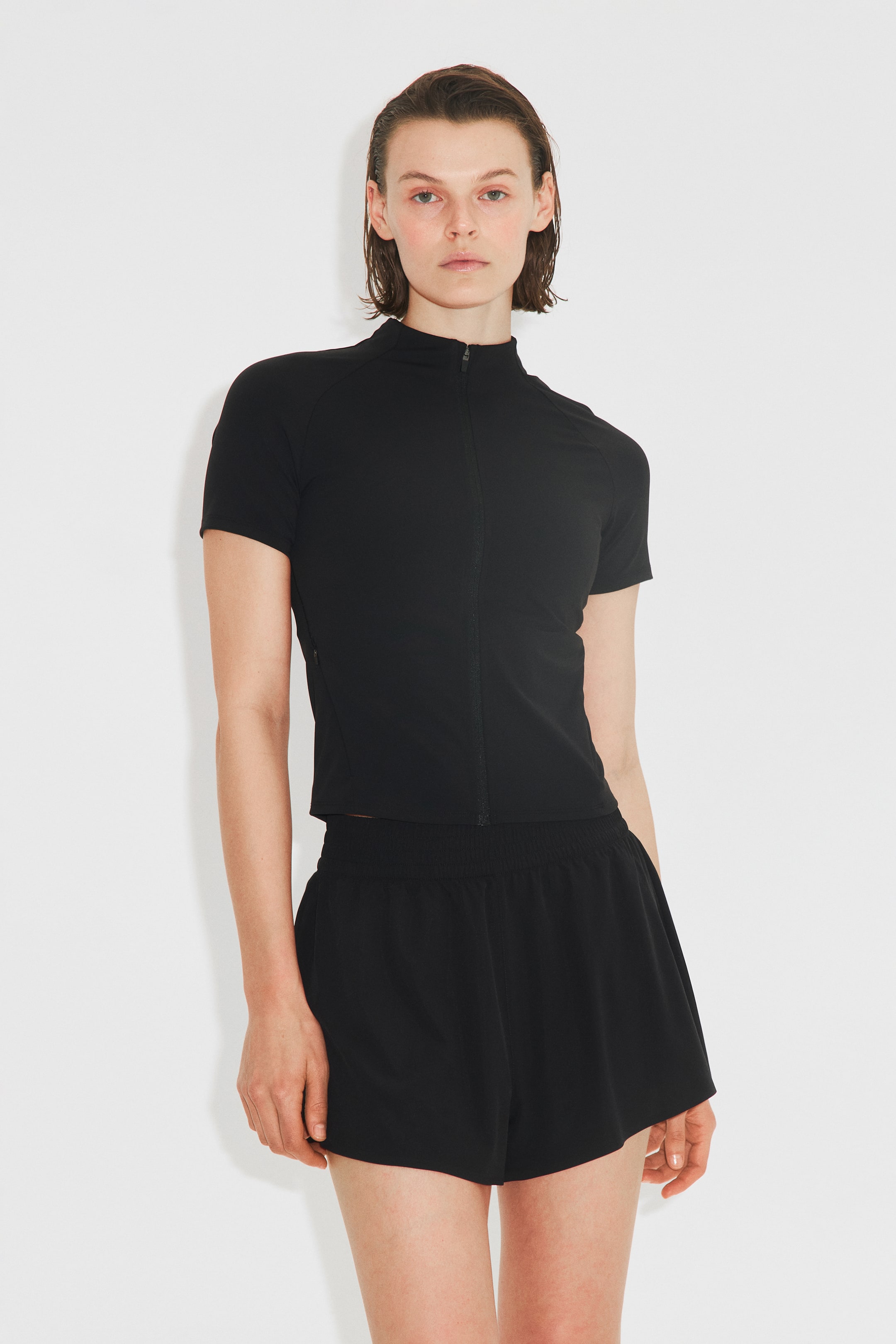 Voir une image plus grande: Une veste noire à manches courtes avec SculptMove, dotée d'un col montant et d'une fermeture éclair à l'avant, est portée par le mannequin, qui porte également un short noir.