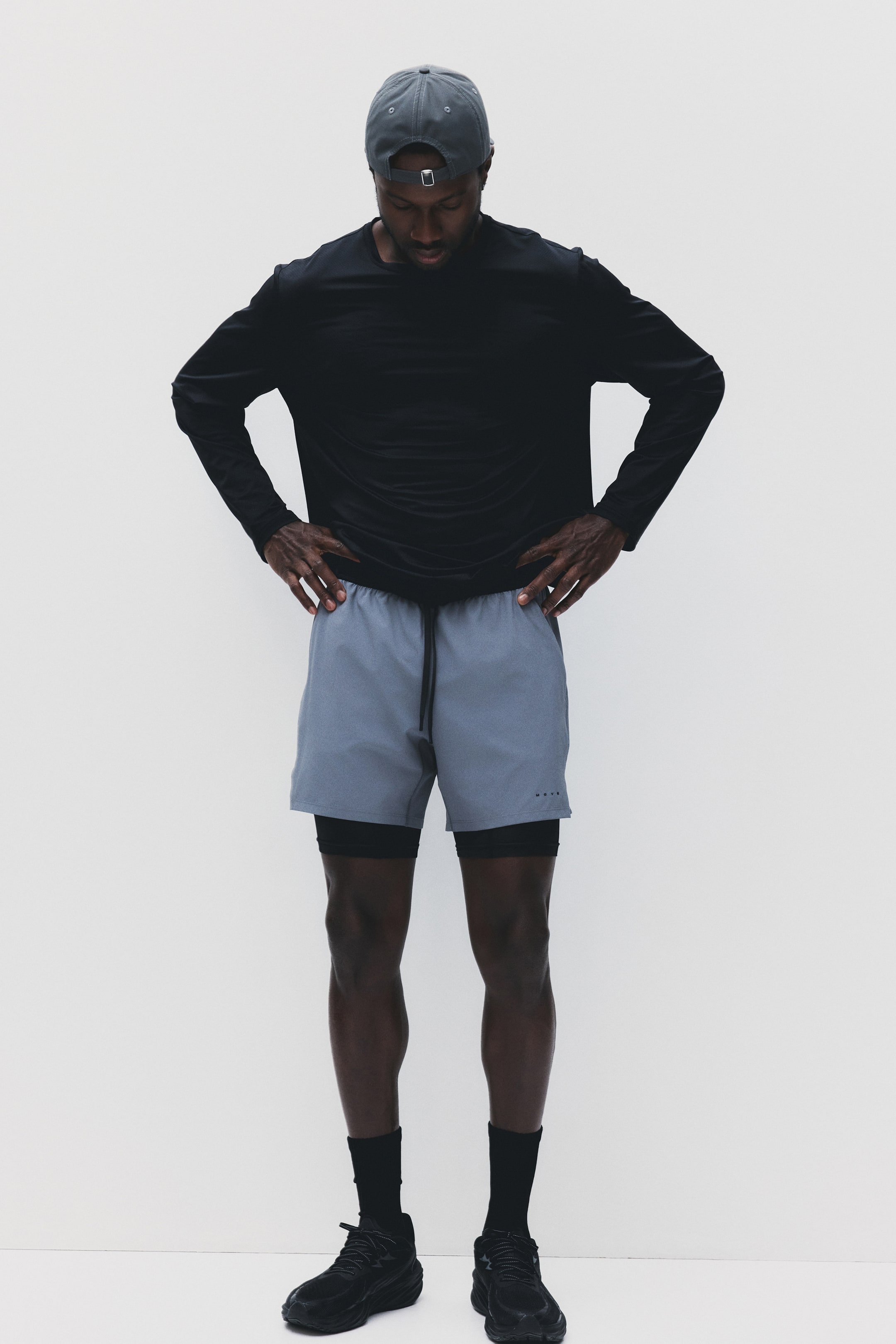 View larger image: A black long-sleeve athletic shirt is being showcased, perfectly matching light grey layered shorts with a black drawstring waist and black compression shorts subtly visible underneath. A dark grey baseball cap sits smartly on the head, while black ankle socks and black athletic shoes complete the sporty look.