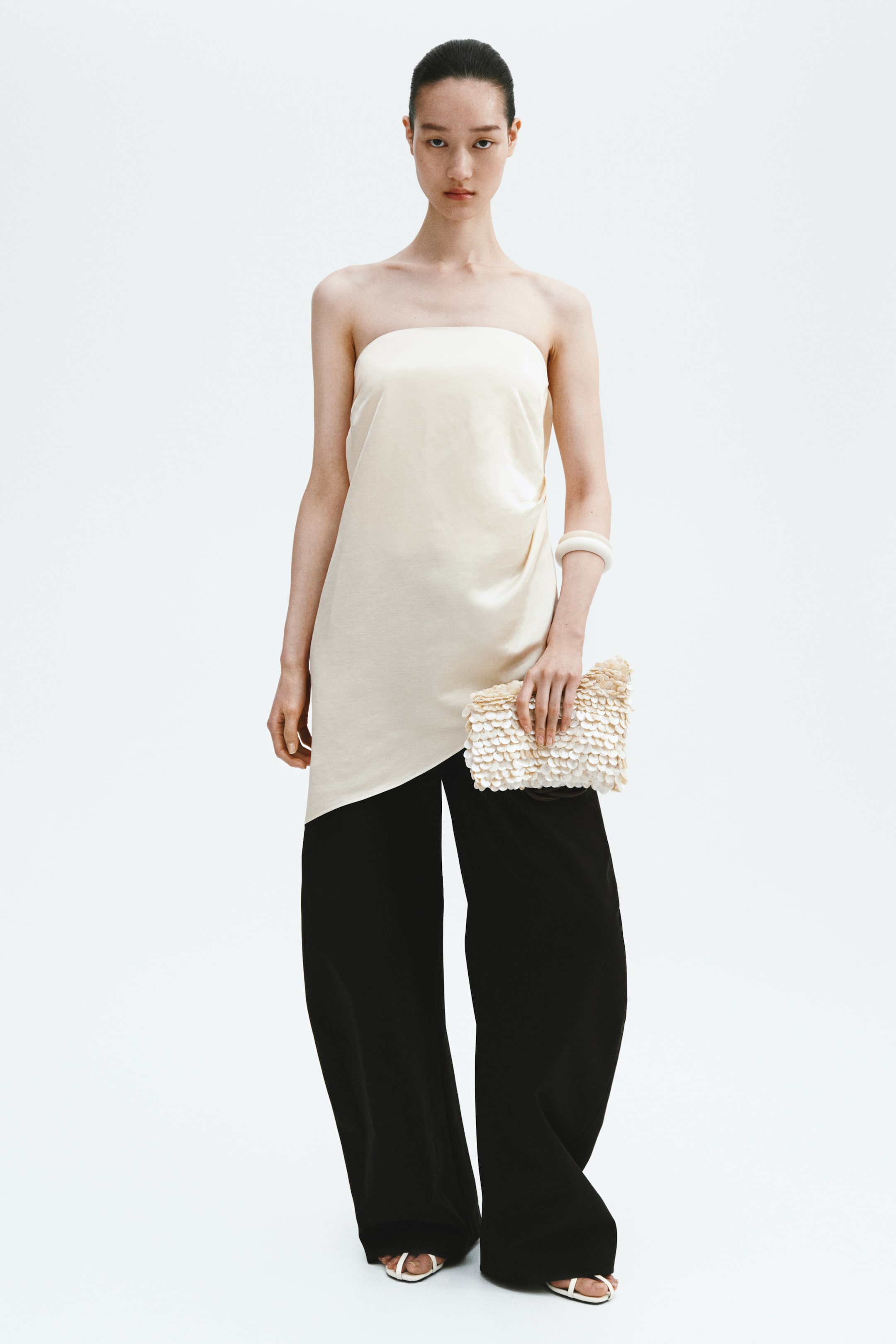 View larger image: The model is showcasing a cream-coloured satin bandeau top, distinguished by pleats on one side of the waist that create a softly draped effect, along with an asymmetrical hem. Complementing the look, the model is wearing black wide-leg trousers and carrying a textured clutch bag.