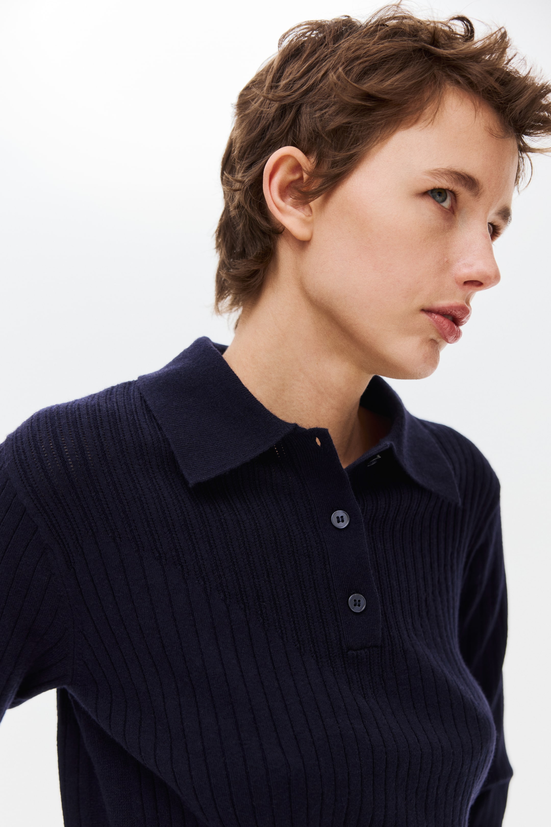 Visa större bild: En mörkblå ribbstickad pikétröja med klassisk krage och en treknappsknäppning med matchande mörka knappar. Plagget presenteras i trekvarts vinkel framifrån och framhäver dess vertikala ribbor.