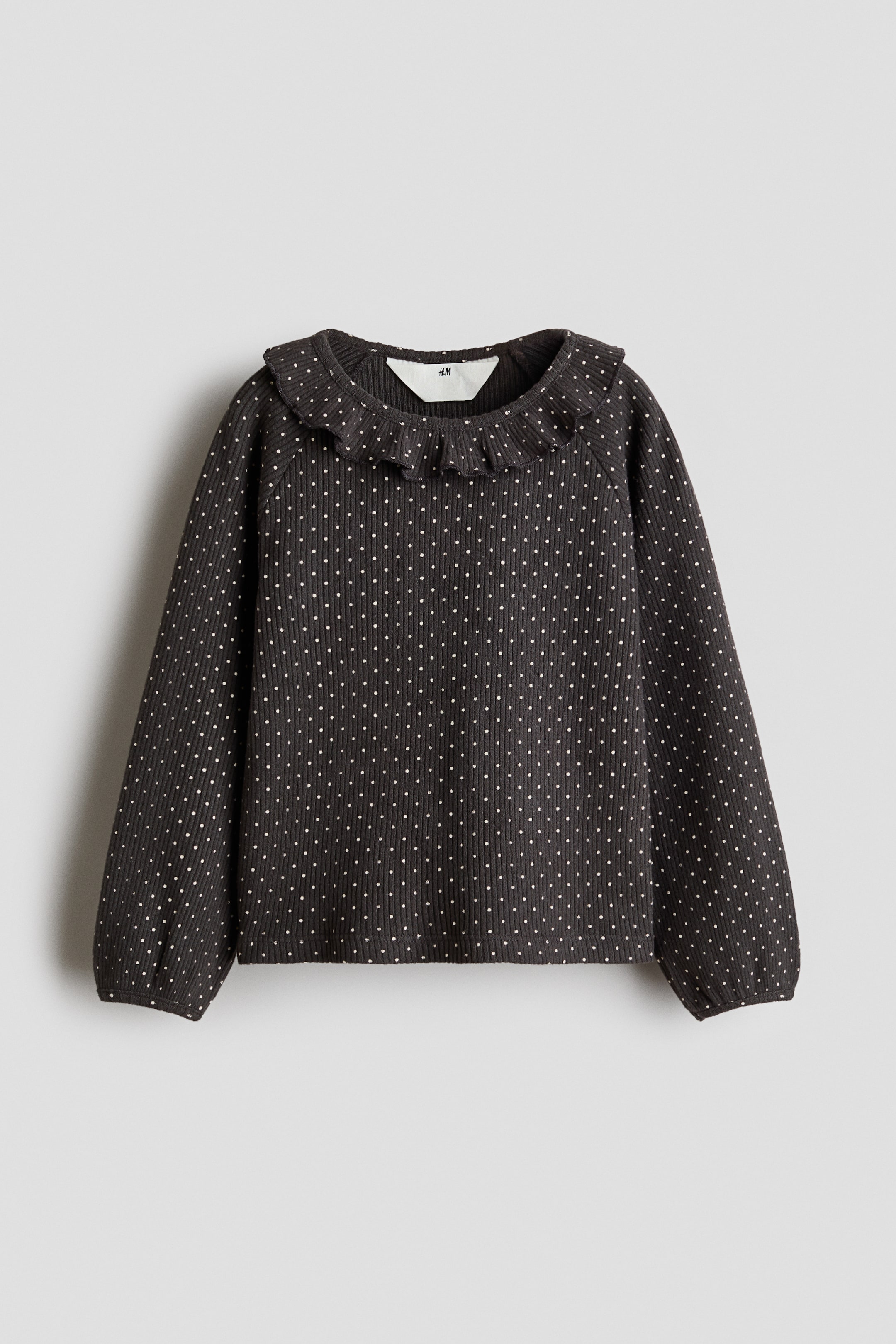View larger image: This dark grey ribbed cotton jersey top with a white polka dot pattern is shown from the front, featuring a round, frill-trimmed collar and long raglan sleeves with narrow binding at the cuffs.