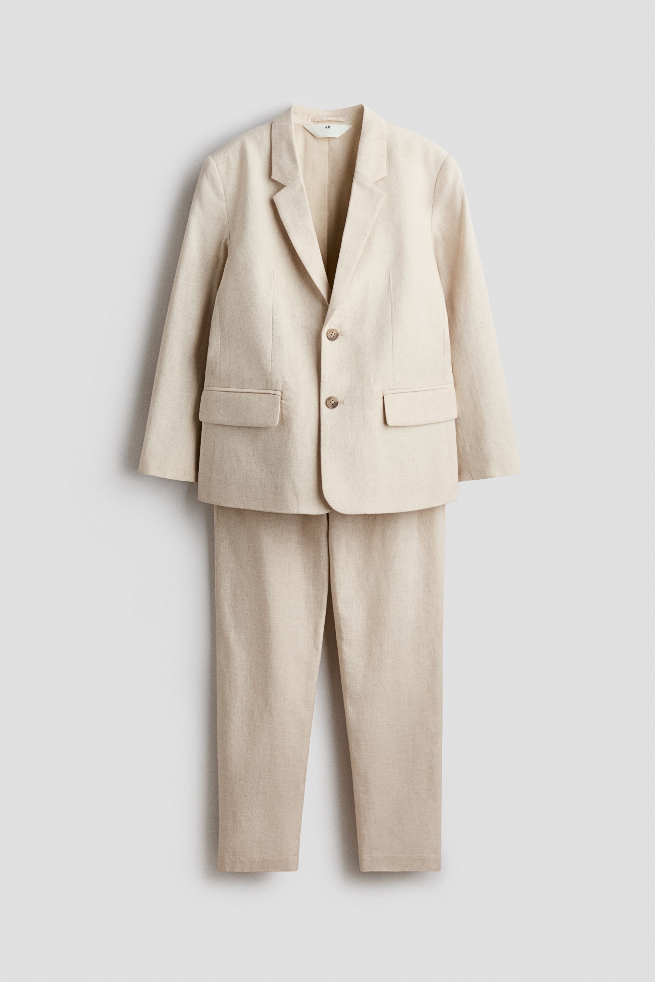 View larger image: A suit crafted from a beige linen and cotton blend showcases its FRONT, with a jacket featuring notch lapels, two front buttons, and flap pockets. The coordinating tailored trousers display a zip fly with a button, diagonal side pockets, and tapered legs.