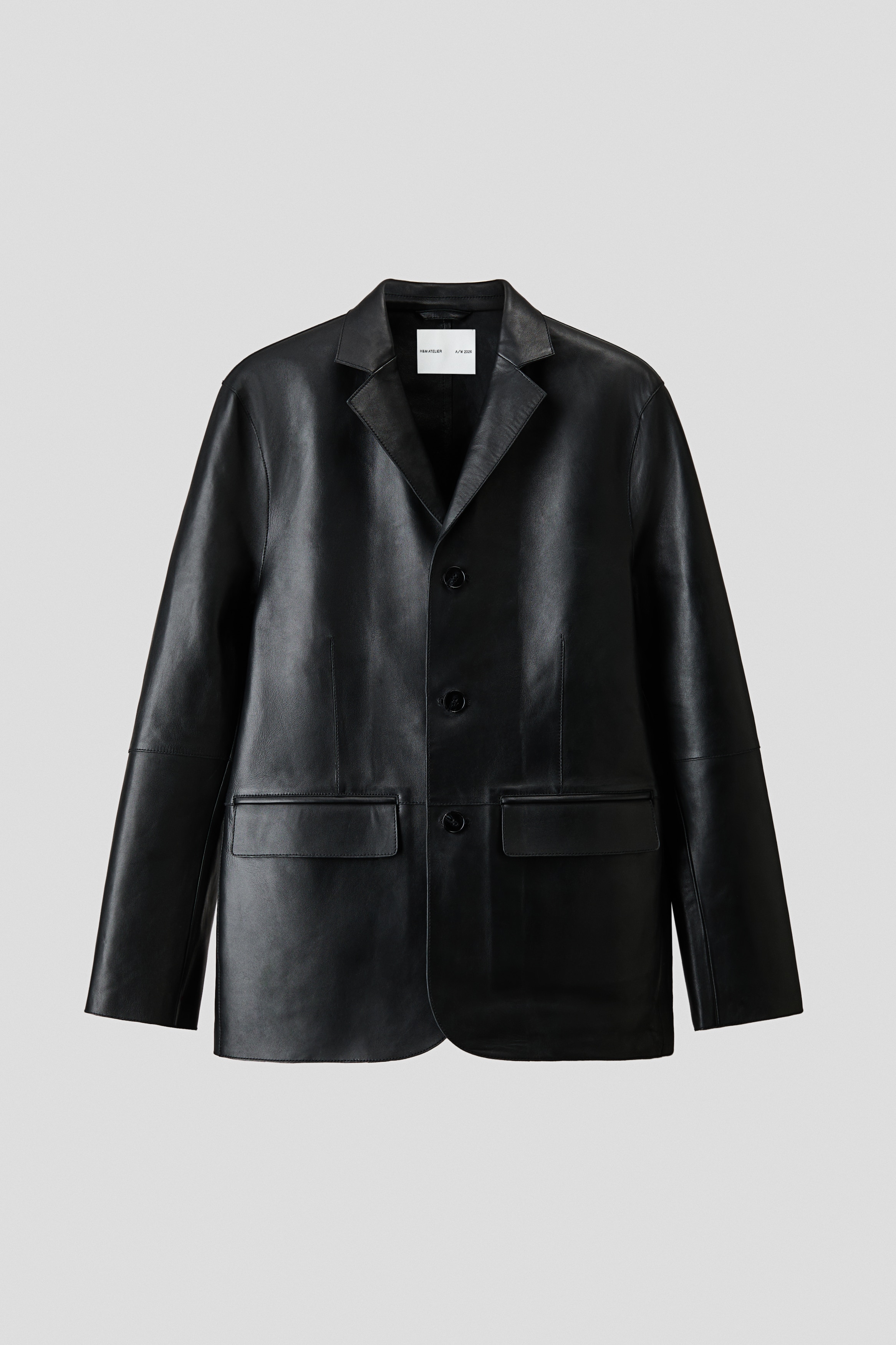 A black leather jacket featuring notch lapels and button closures is being worn by the model. The jacket is styled as a layer over a white collared shirt with a striped tie and paired with dark blue jeans.