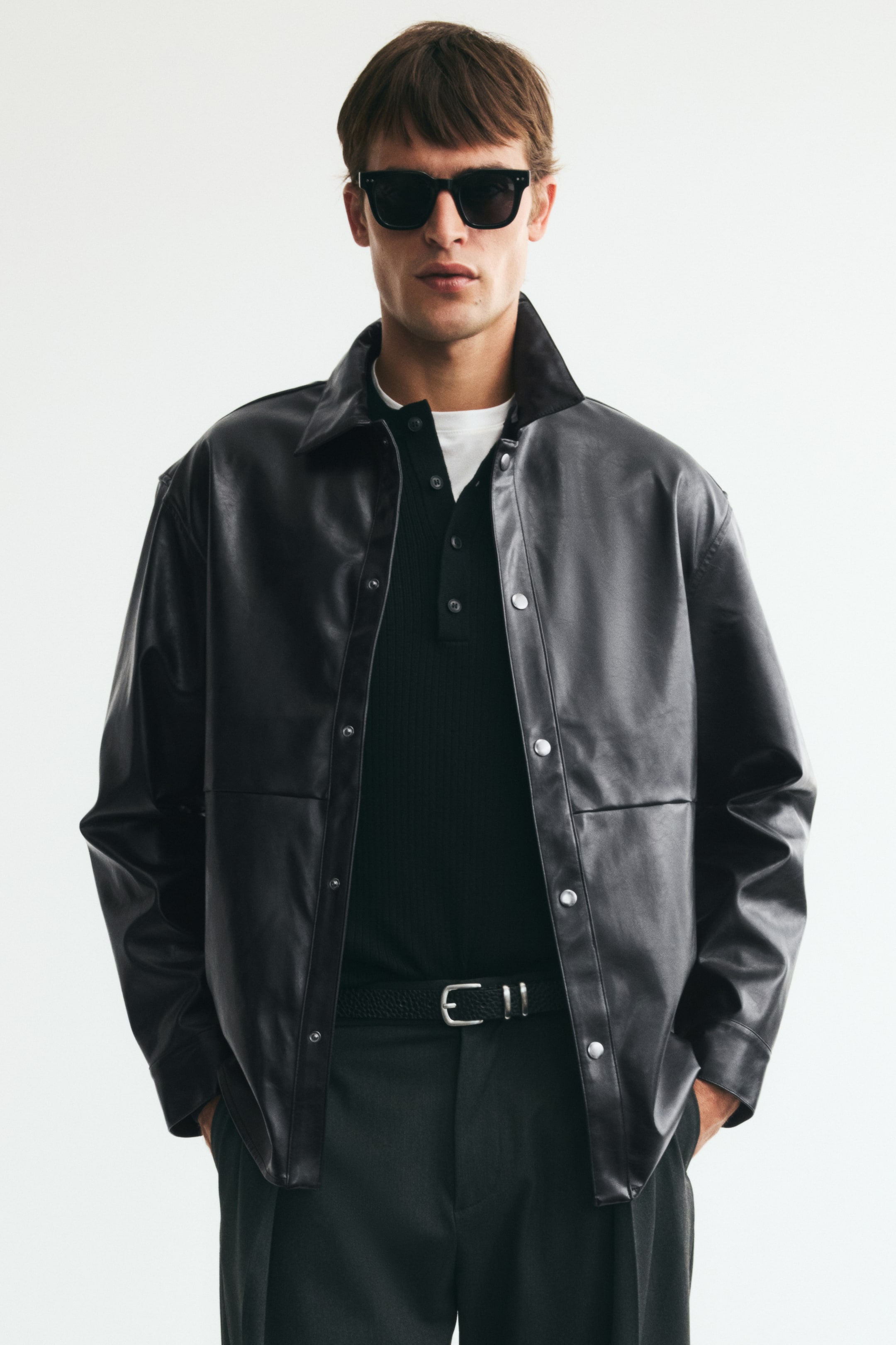 View larger image: The model is wearing an overshirt in coated fabric. It features a collar, press studs down the front, a front yoke, press-stud cuffs, and a gently rounded hem. The model is wearing the overshirt unfastened over a white T-shirt and a black ribbed polo shirt.