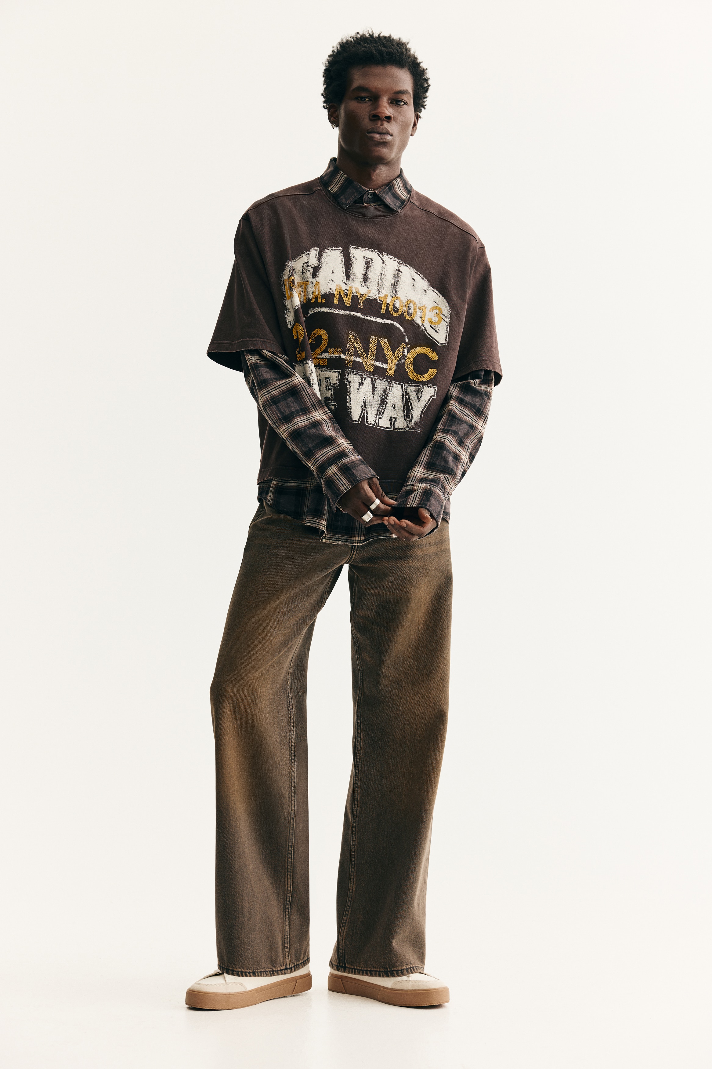 A dark brown, short-sleeved t-shirt is being presented from the front, featuring a prominent distressed graphic print. The print displays layered text, including "LEADING" and "THE WAY" in white, and "B'KLYN NY 10013" and "212-NYC" in gold, with the "212-NYC" text enclosed in an outline, all set against the t-shirt's crew neck and raw-edge hem.