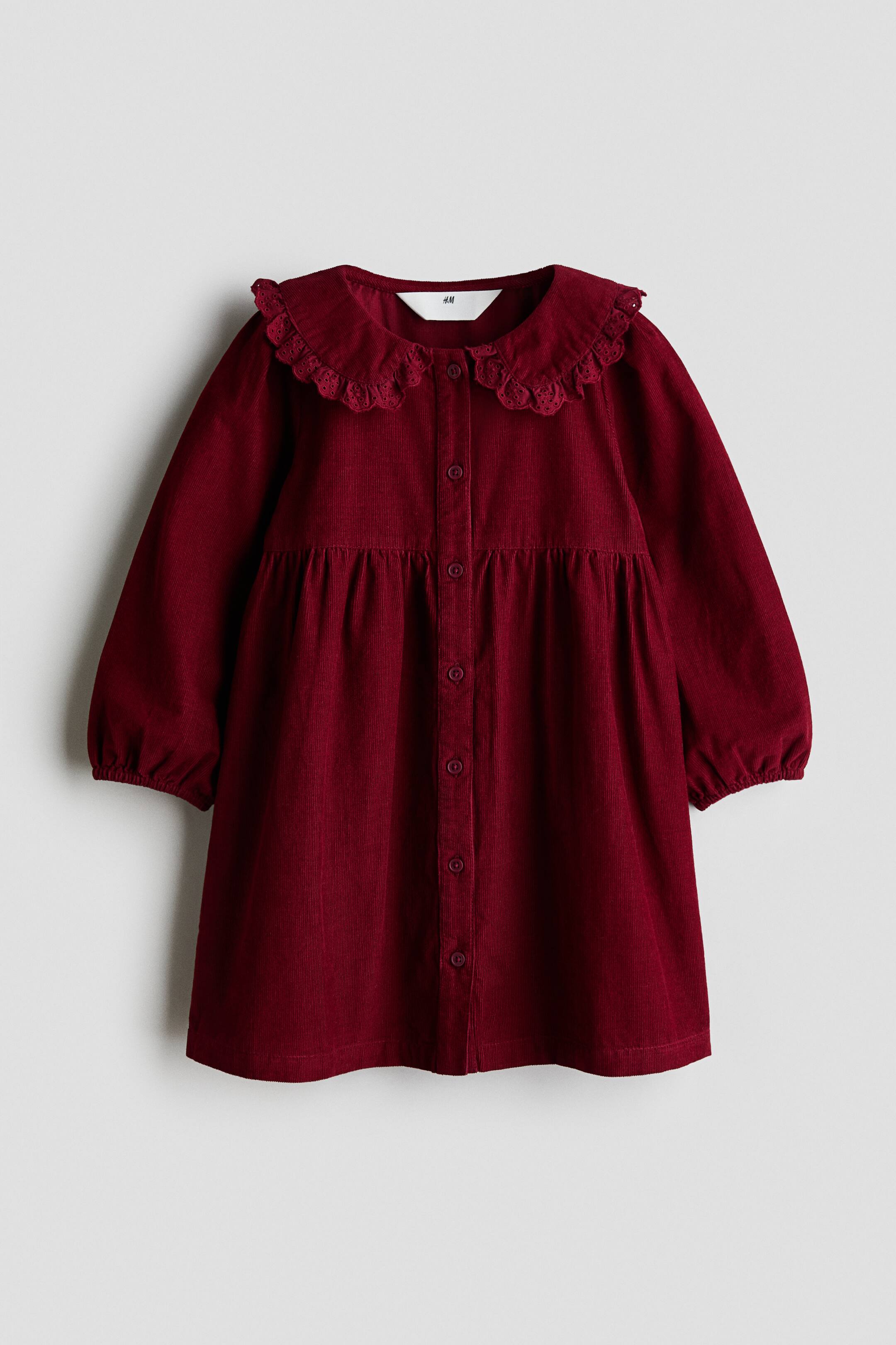 Agrandir l'image: Une robe en velours côtelé de coton rouge foncé est présentée de face, avec un col Claudine arrondi orné d’une bordure festonnée en broderie anglaise. Le modèle comprend une patte de boutonnage sur toute la longueur à l’avant, des manches longues avec poignets élastiqués et une couture froncée à la taille.