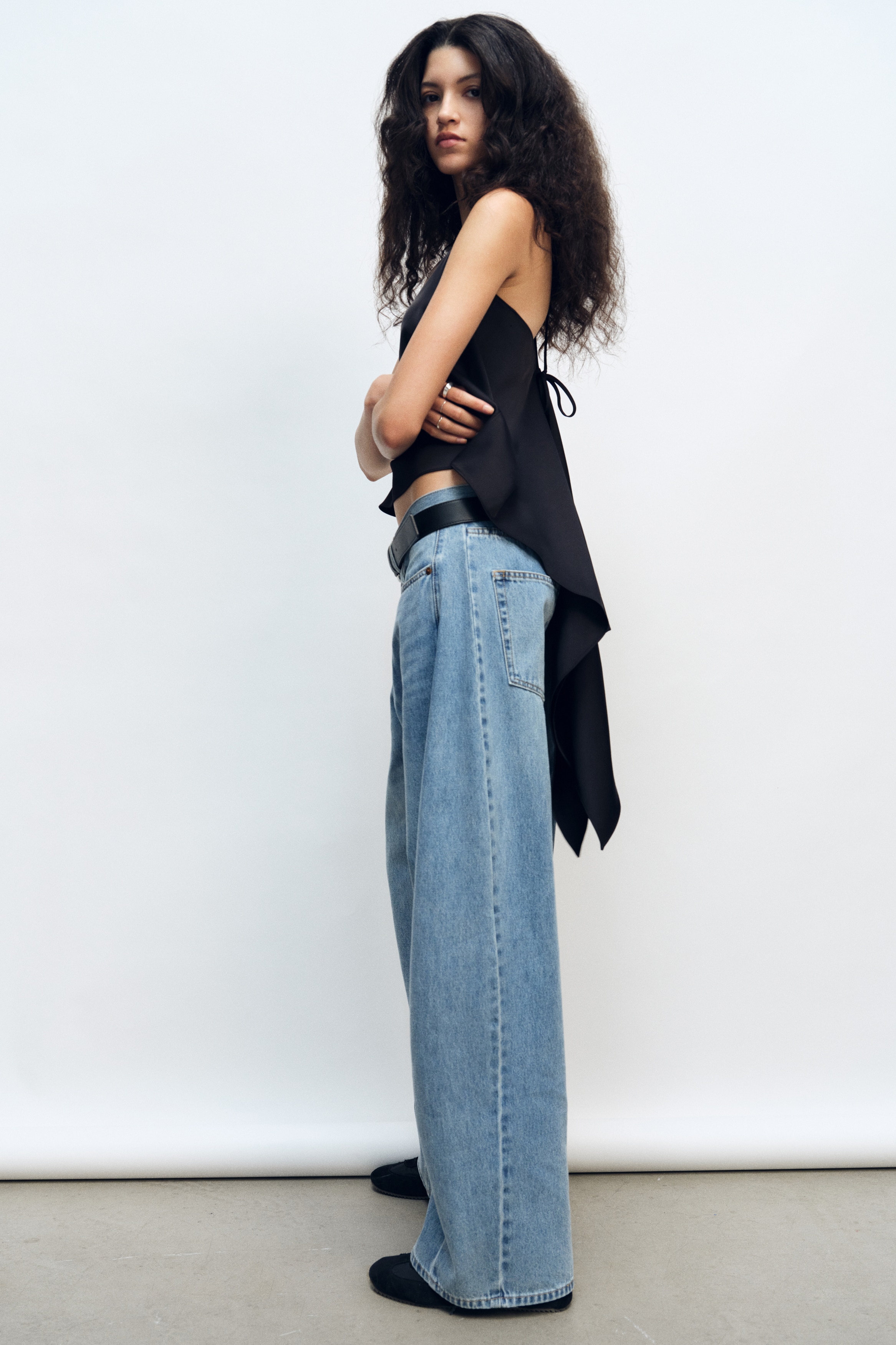 Een zwarte camisole-top hangt met de voorkant naar voren en laat smalle spaghettibandjes zien die over de borst kruisen. De soepele stof valt in een gelaagd silhouet, met lange, asymmetrische banen die onder de rechte zoomlijn naar beneden vloeien.