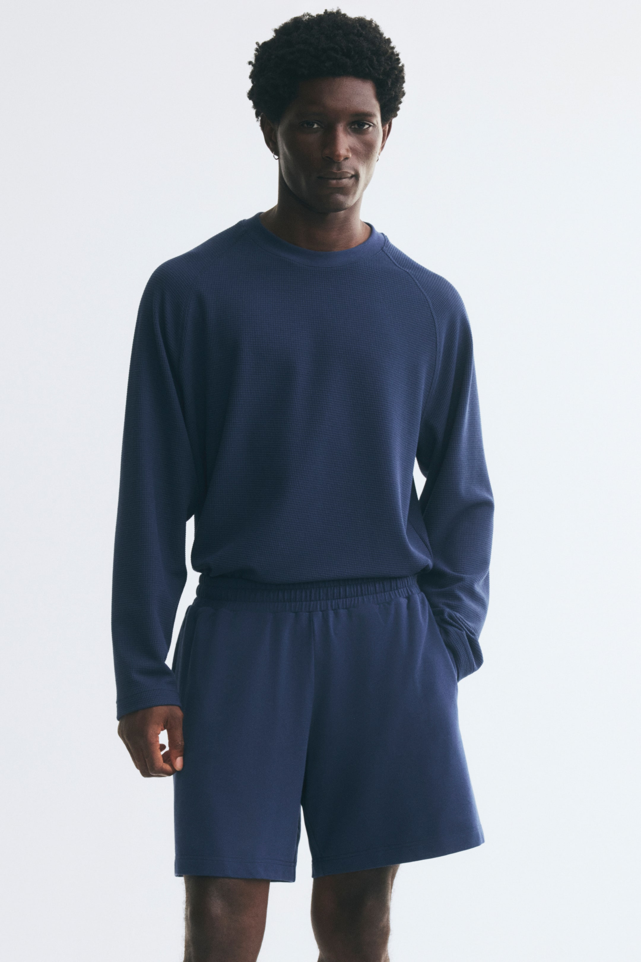 View larger image: Blue sweat shorts with an elasticated waistband and a regular fit are worn by the model, who is standing with one hand in a side pocket. The shorts are styled with a matching blue textured long-sleeve top.
