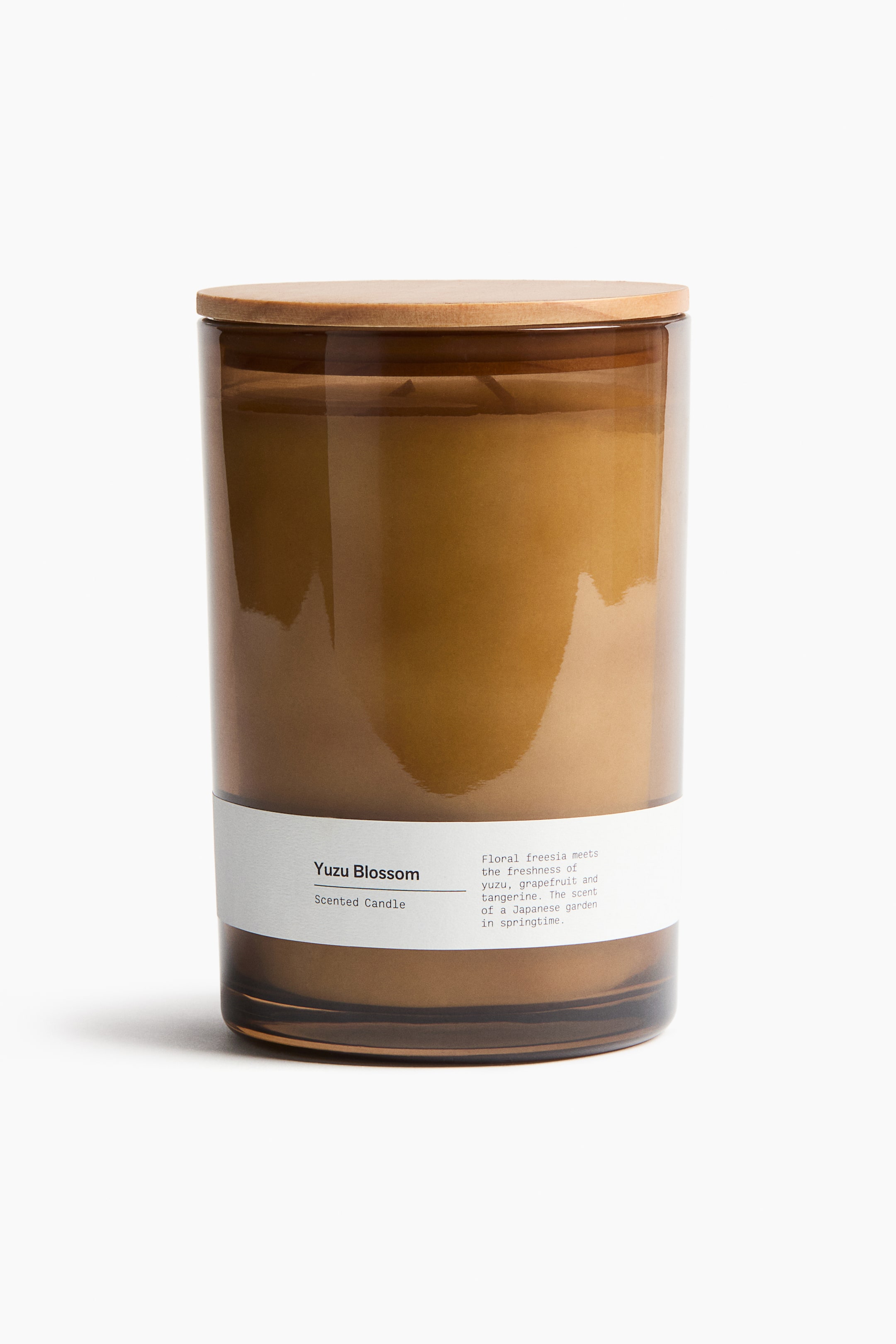 View larger image: A scented candle is housed in a cylindrical, amber-tinted glass holder, topped with a light brown pinewood lid. A white label is wrapping around the front, displaying "Yuzu Blossom Scented Candle" along with a description of floral freesia, yuzu, grapefruit, and tangerine notes.