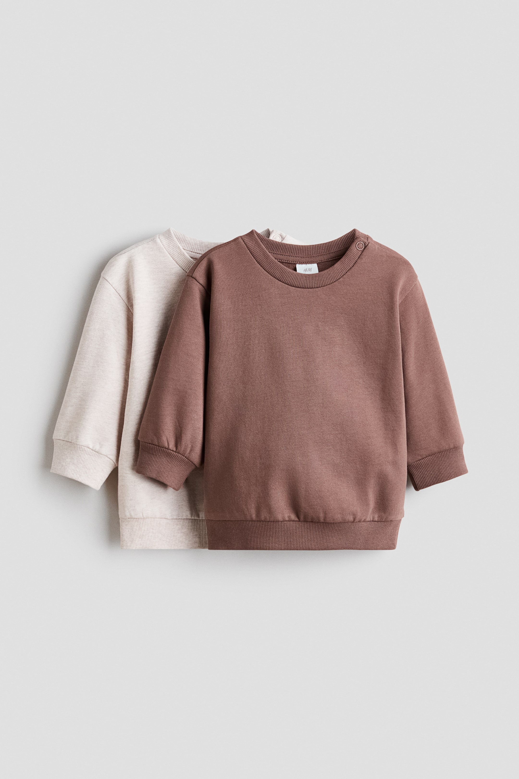 View larger image: Two children's sweatshirts, with a brown one at the forefront and a light beige one positioned behind, both showcasing long sleeves, ribbed neck, cuffs, and hem. The brown sweatshirt further features a snap fastener on its left shoulder.