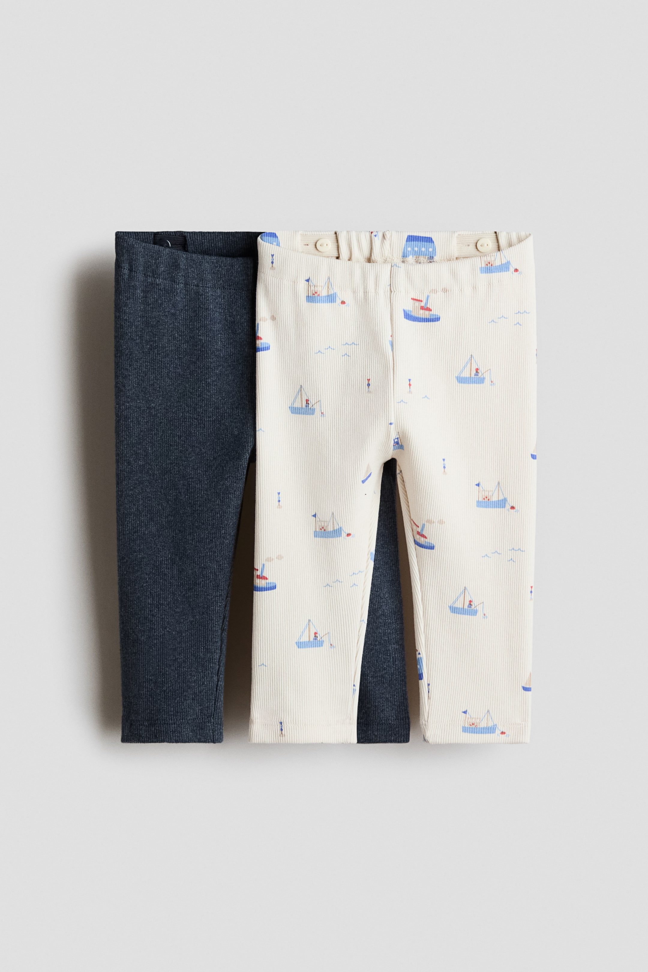 Visualizza immagine più grande: Due paia di leggings a costine jersey H&M Adorables sono esposti frontalmente, stesi piatti. Un paio è di un blu scuro uniforme, e l'altro è bianco sporco con un motivo ripetuto di barche a vela blu e figure, entrambi con una fascia elastica in vita con bottoni regolabili.