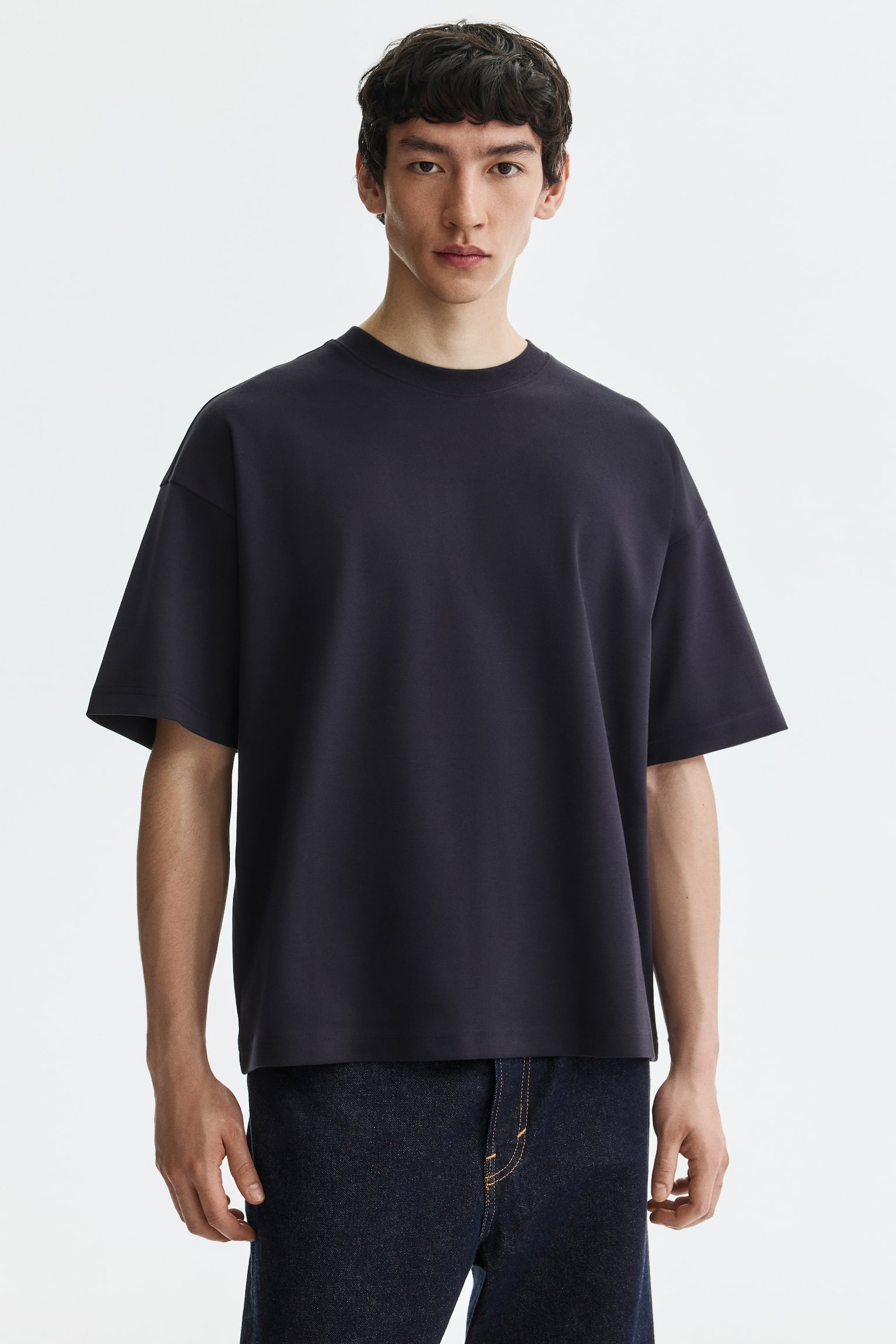 A dark grey crew neck t-shirt is being worn frontally, showcasing its smooth fabric, relaxed fit, and short, wide sleeves. - 1