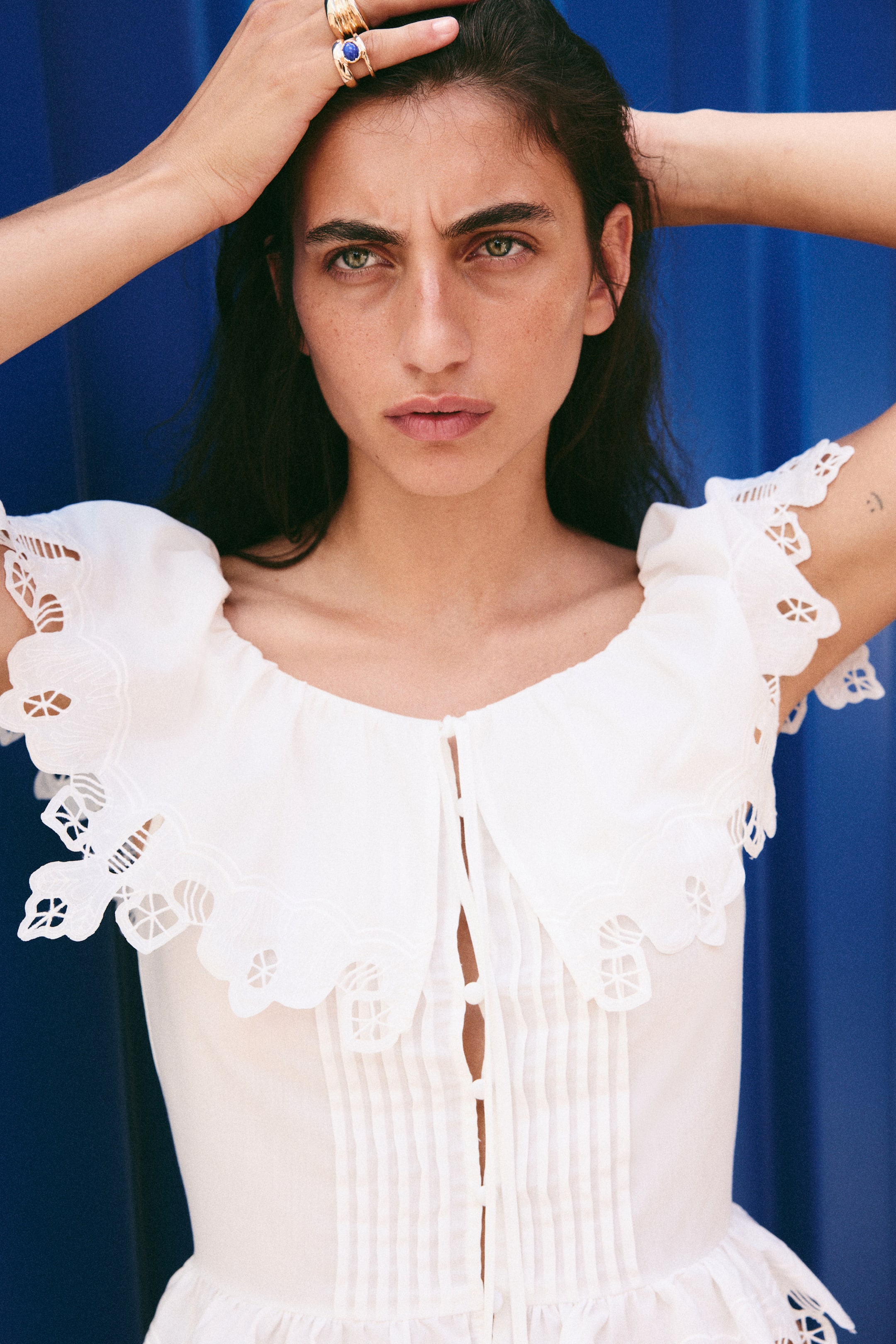View larger image: The model is wearing a white fitted blouse with broderie anglaise and scalloped edges. It features a flounced ballerina neckline with a drawstring creating a keyhole opening, and delicate pintucks with covered buttons down the front, with some buttons undone. A wide frill trim is visible at the hem.