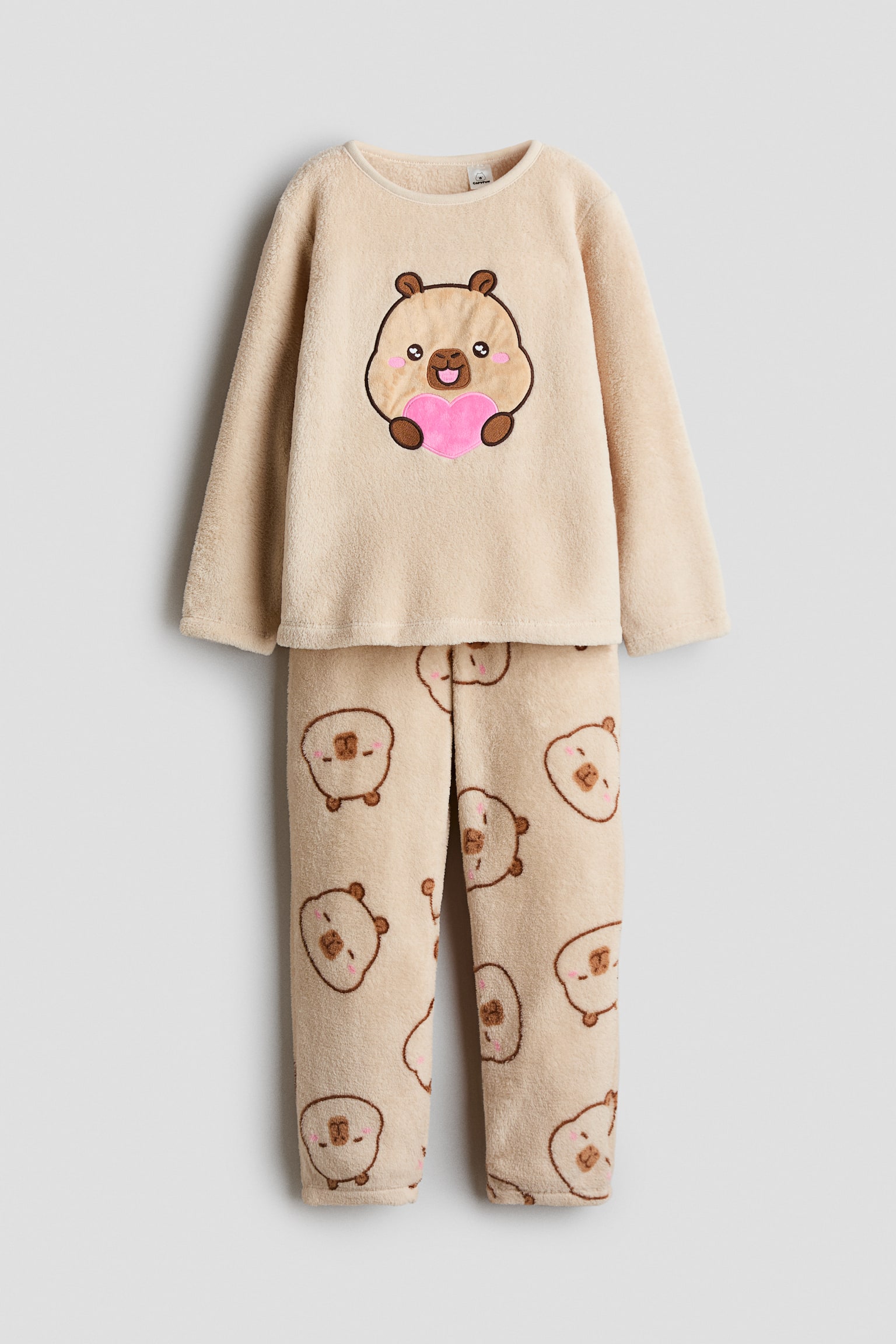 Un ensemble de pyjama en velours beige est prÃ©sentÃ© de face, comprenant un haut Ã  manches longues avec un col rond et un grand capybara de dessin animÃ© tenant un cÅur rose sur la poitrine. Le pantalon assorti prÃ©sente un imprimÃ© all-over de petits visages de capybara.