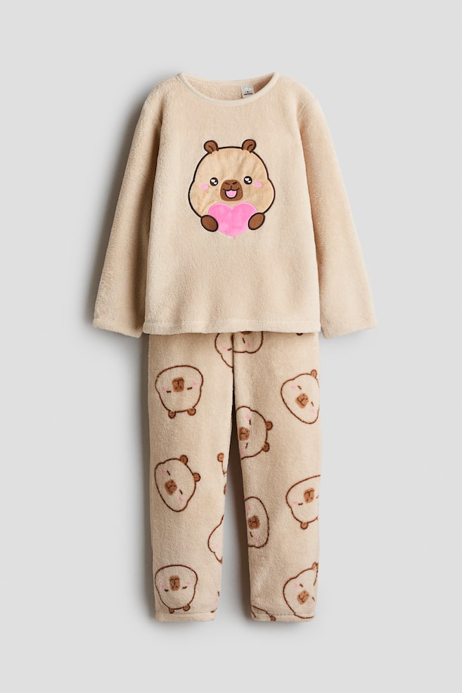 Ein beiges Velours-Pyjamaset ist von vorne zu sehen, bestehend aus einem langÃ¤rmeligen Oberteil mit Rundhalsausschnitt und einem groÃen Cartoon-Kapibara, das ein rosa Herz auf der Brust hÃ¤lt. Die dazu passende Hose zeigt ein allover-Print aus kleineren Kapibara-Gesichtern. - 1