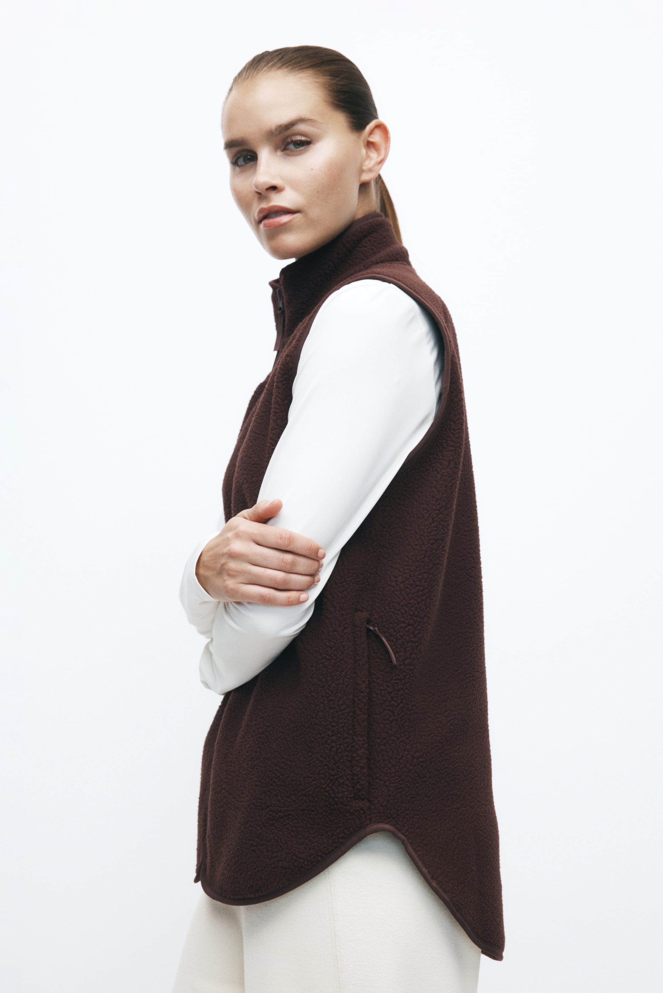 Größeres Bild ansehen: Eine dunkelbraune Teddy-Fleece-Activewear-Weste wird vom Model über einem langärmligen weißen Oberteil und cremefarbenen Hosen getragen. Die Weste verfügt über einen hohen Kragen, eine seitliche Reißverschlusstasche und einen abgerundeten Saum, der hinten länger geschnitten ist.