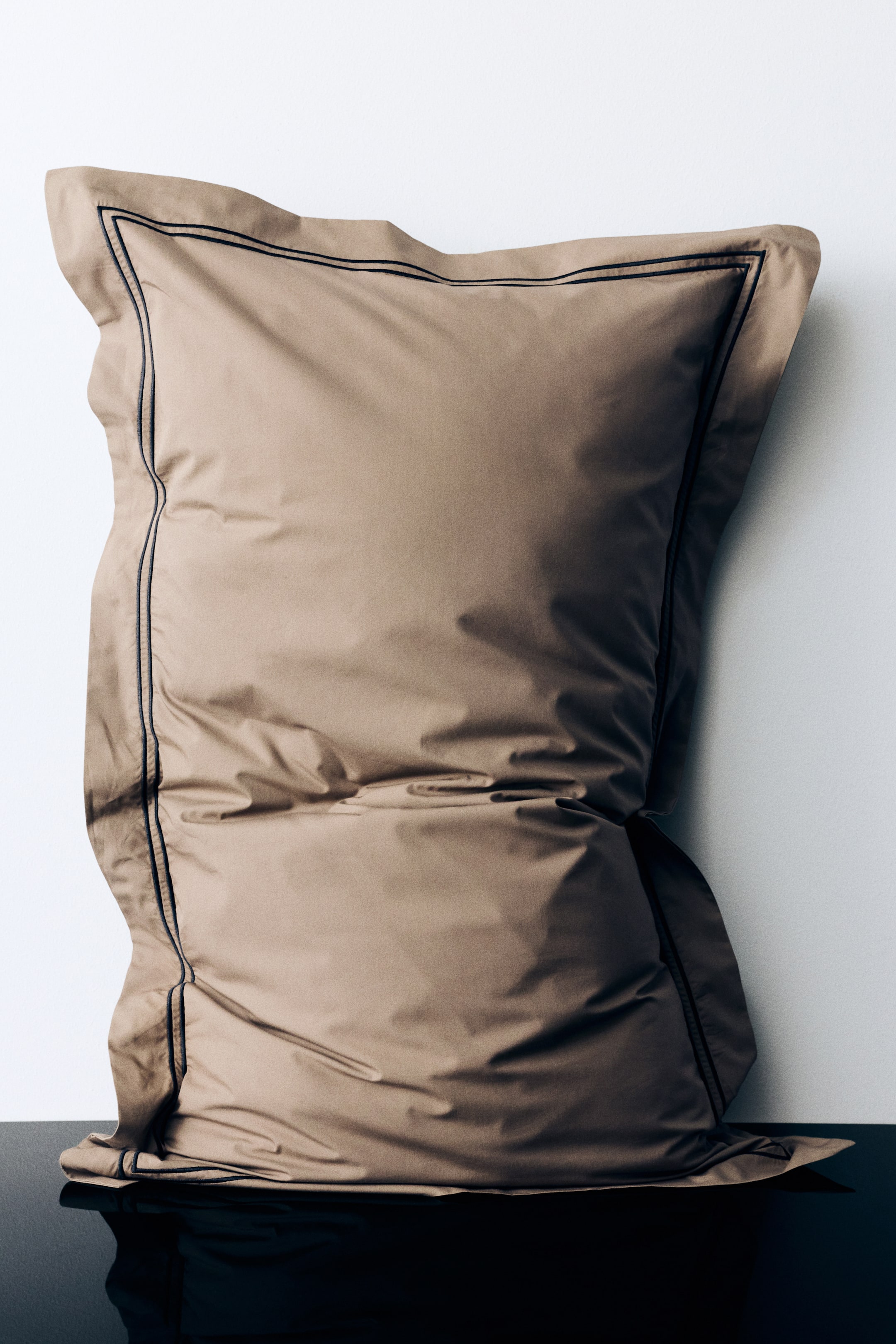 View larger image: A taupe percale cotton pillowcase stands upright in a front view, showing its filled shape with a wide border around the sides. The pillowcase features a thin, double-lined dark embroidery framing the inner edge of the border.