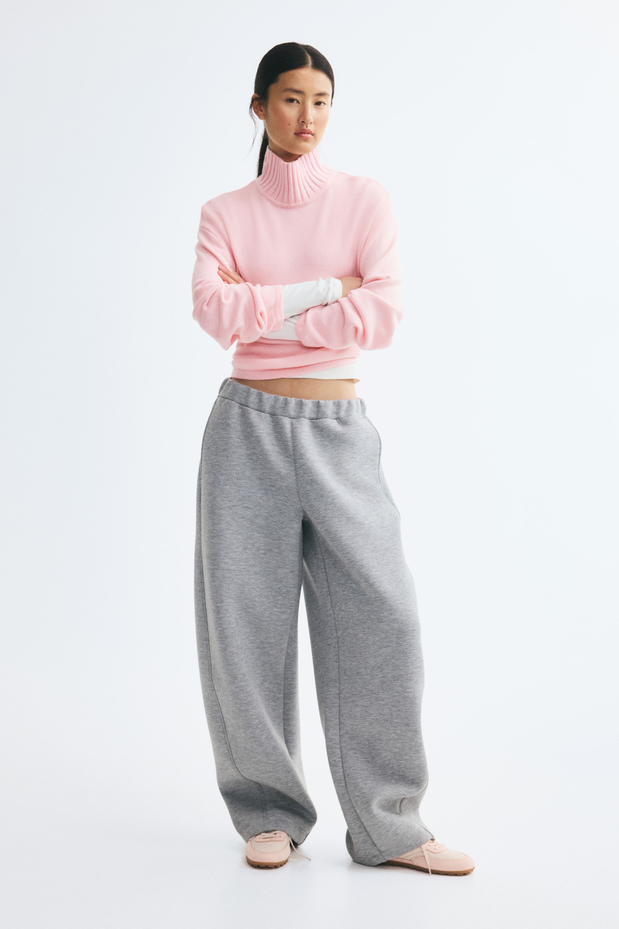 View larger image: Facing front, these wide-leg sweatpants in heather grey showcase a relaxed, oversized fit with a gathered elastic waistband. The trousers pool slightly at the hem over pink sneakers and are paired with a cropped pink turtleneck sweater.