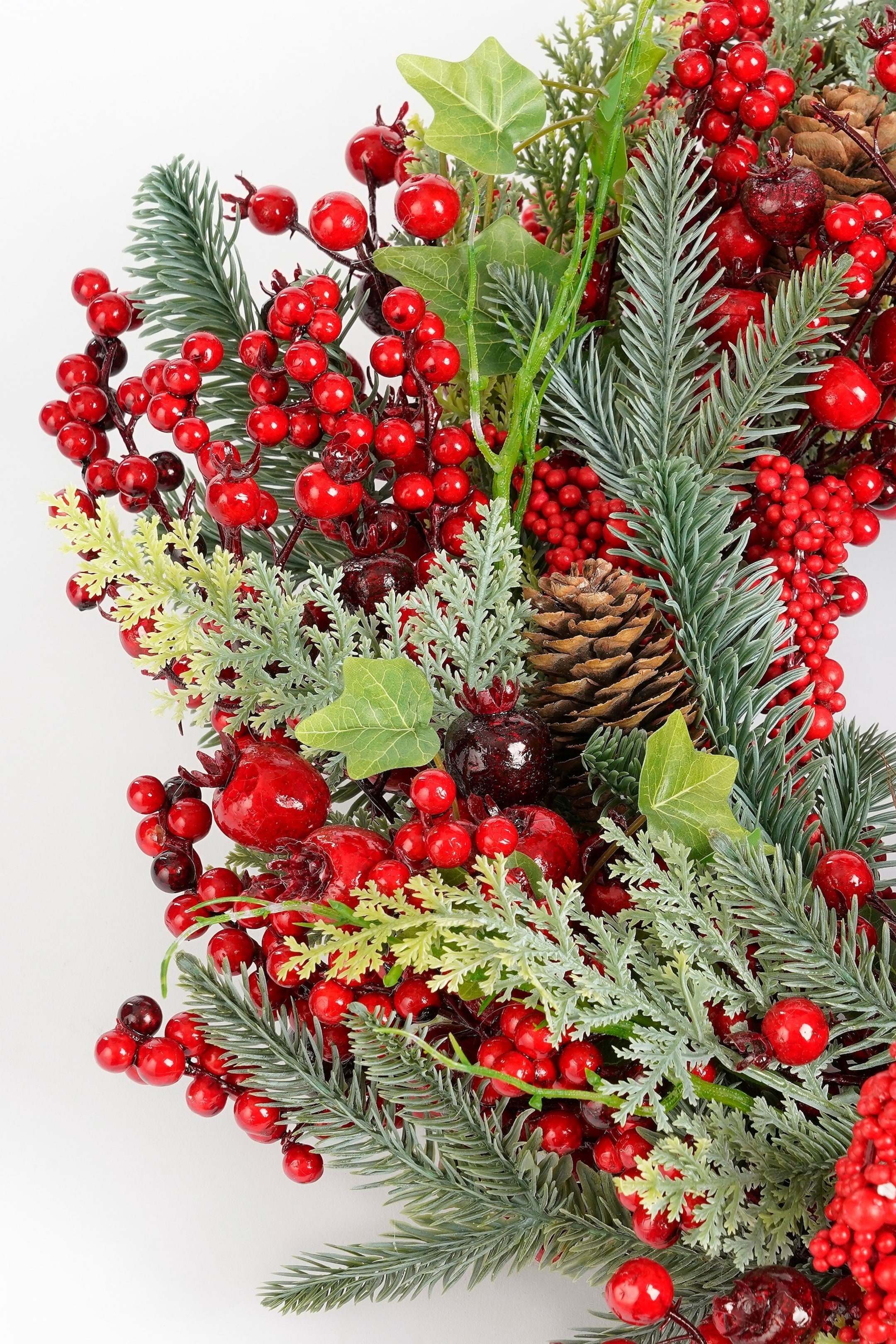 Grotere afbeelding bekijken: Een kunstmatige schikking toont dichte clusters van glanzend rode bessen, afgewisseld met groene dennen takken en lichter groen, veerachtig loof. Een bruine dennenappel ligt tussen de elementen ingebed, en heeft ook donkerrode, bolvormige accenten.