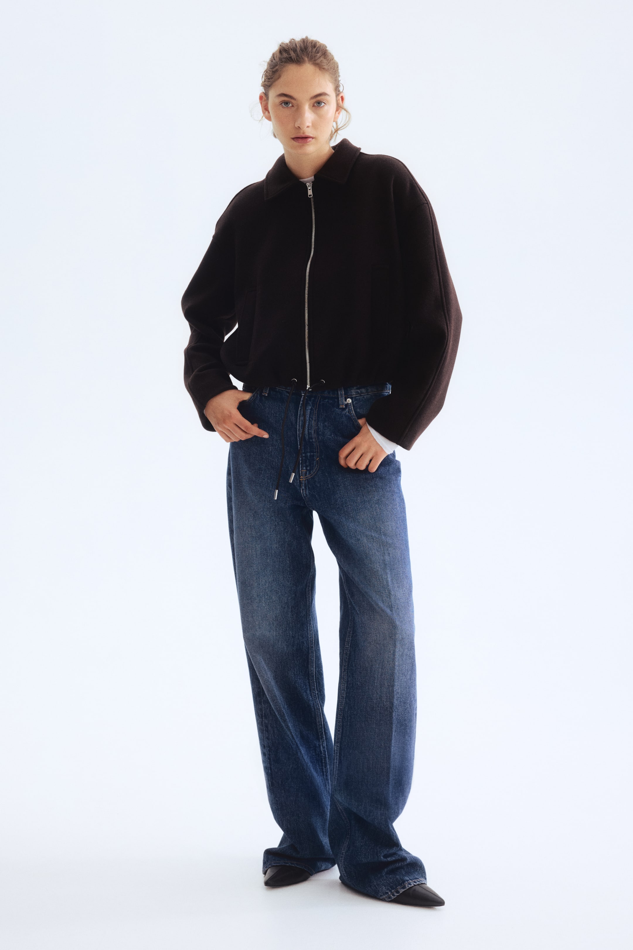 Agrandir l'image: Une veste courte marron foncé avec un col pointu, une fermeture éclair complète de couleur argentée et un ourlet à cordon de serrage, tandis que des poignets de chemise blancs s'étendent de ses manches amples. Un jean bleu en denim à coupe large et ample présente une taille haute avec une braguette à boutons visible et un détail de cordon noir.