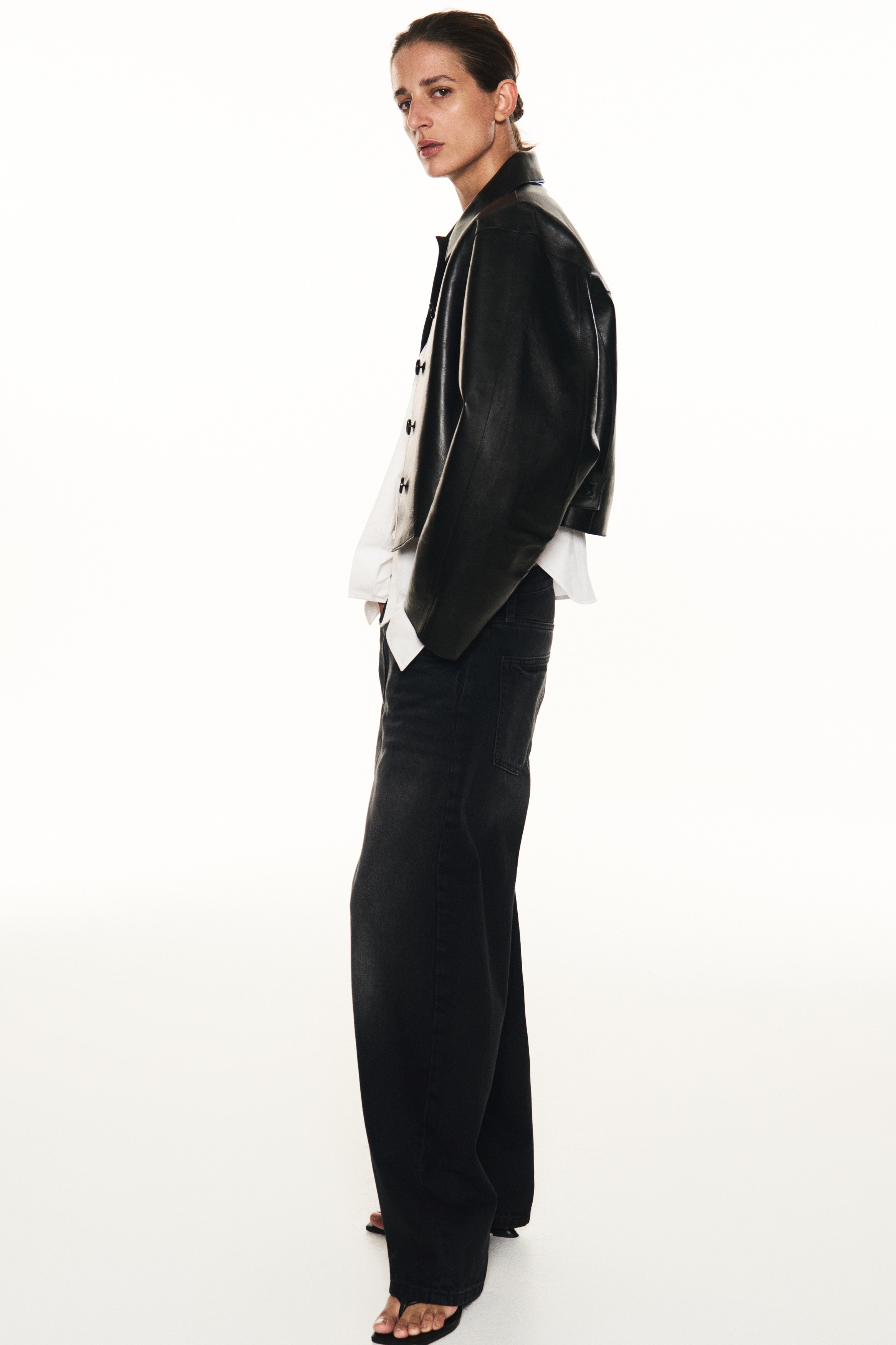 A pair of faded black denim jeans is shown from the front, showcasing a regular waist and a curved, voluminous leg silhouette that tapers towards the hems. The design features a classic five-pocket layout, a button fastening, and horizontal dart seams across the knees.