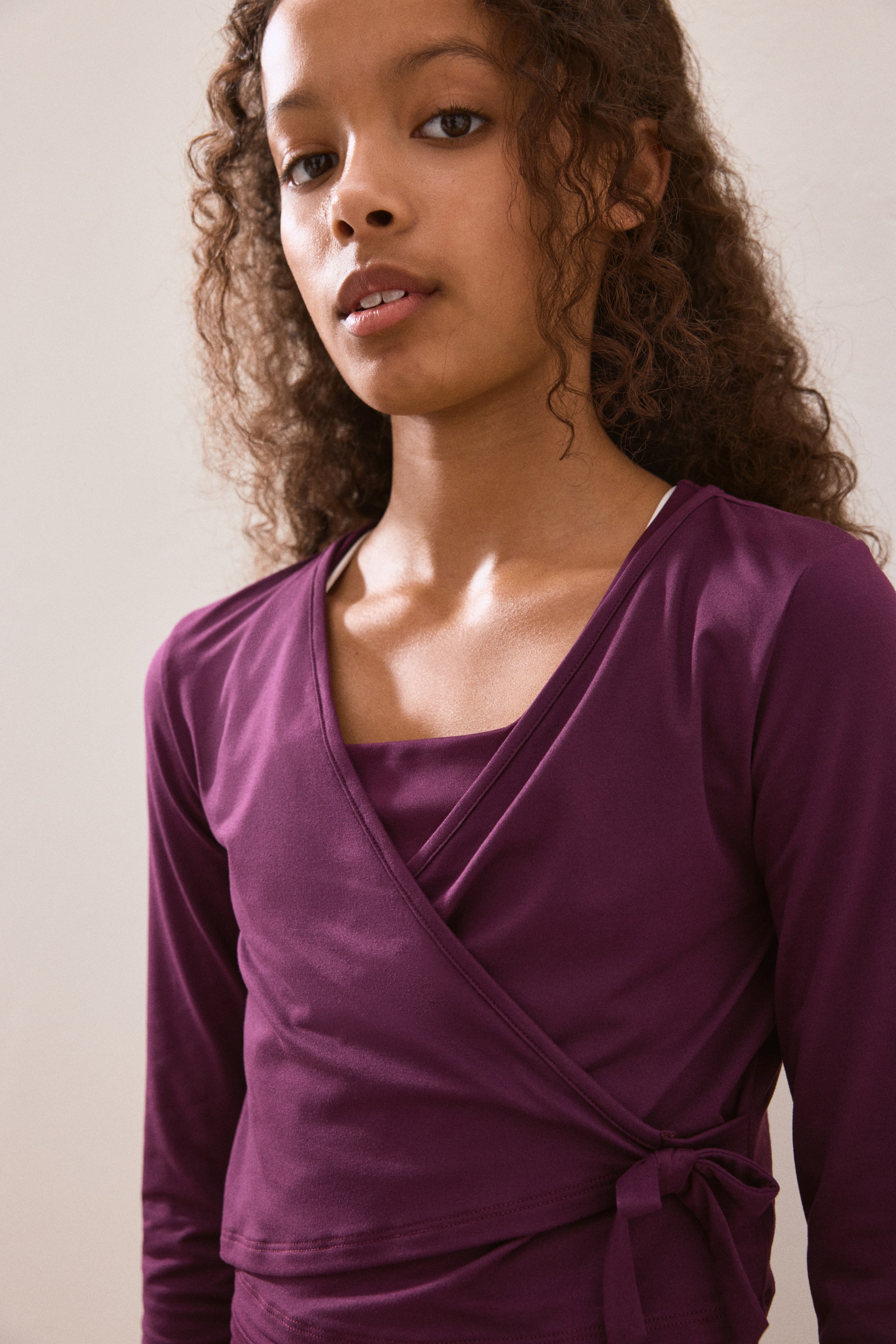 View larger image: "A plum-coloured wrap top is being worn, featuring long sleeves and a V-neckline formed by the overlapping front panels. It is tying on the side, and a lighter garment is layering underneath at the neckline."