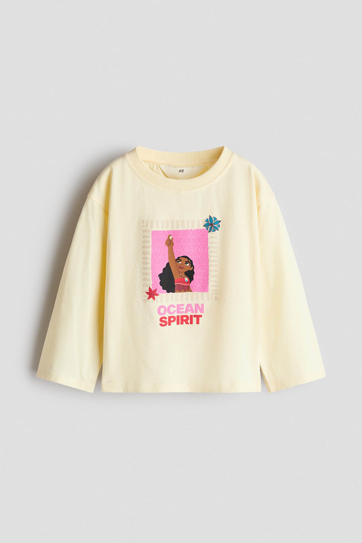 Uma t-shirt de manga comprida em amarelo-claro apresenta uma vista frontal, com decote redondo e acabamento canelado estreito, além de ombros descaÃdos. A frente da camisola apresenta uma impressão gráfica colorida da personagem Moana dentro de um quadrado rosa, com a frase "OCEAN SPIRIT" impressa abaixo em letras rosa e vermelhas.