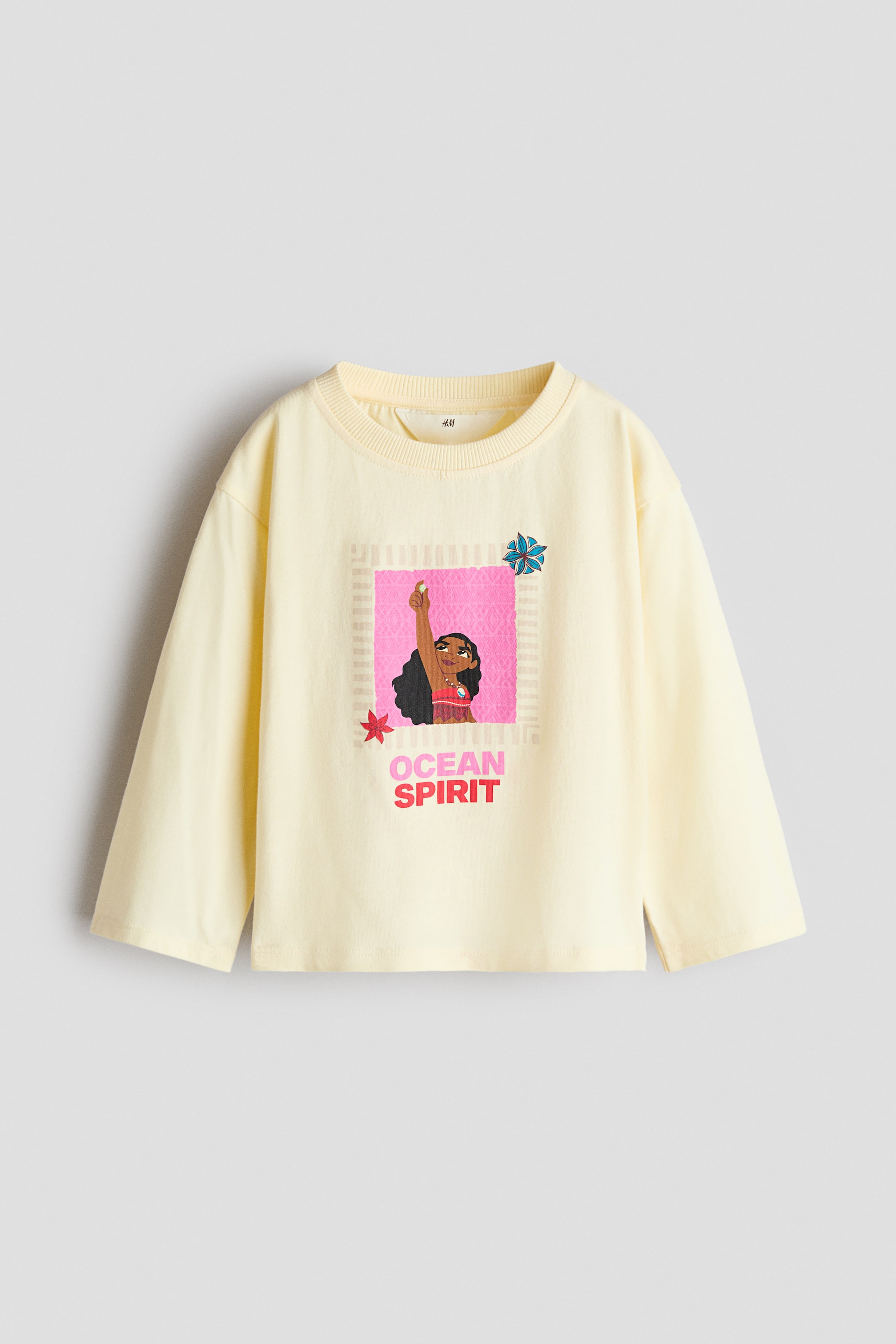 Agrandir l'image: Un t-shirt à manches longues jaune pâle est présenté de face, avec un col rond doté d’une fine bordure côtelée et des épaules tombantes. L’avant du t-shirt arbore un imprimé graphique coloré du personnage de Moana à l’intérieur d’un carré rose, avec la phrase « OCEAN SPIRIT » imprimée en dessous en lettres roses et rouges.