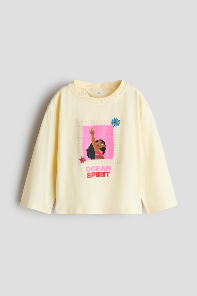 A pale yellow long-sleeve T-shirt is shown in a front-facing view, featuring a round neckline with narrow ribbed trim and dropped shoulders. The front of the shirt features a colourful graphic print of the character Moana inside a pink square, with the phrase "OCEAN SPIRIT" printed below in pink and red letters. - 1