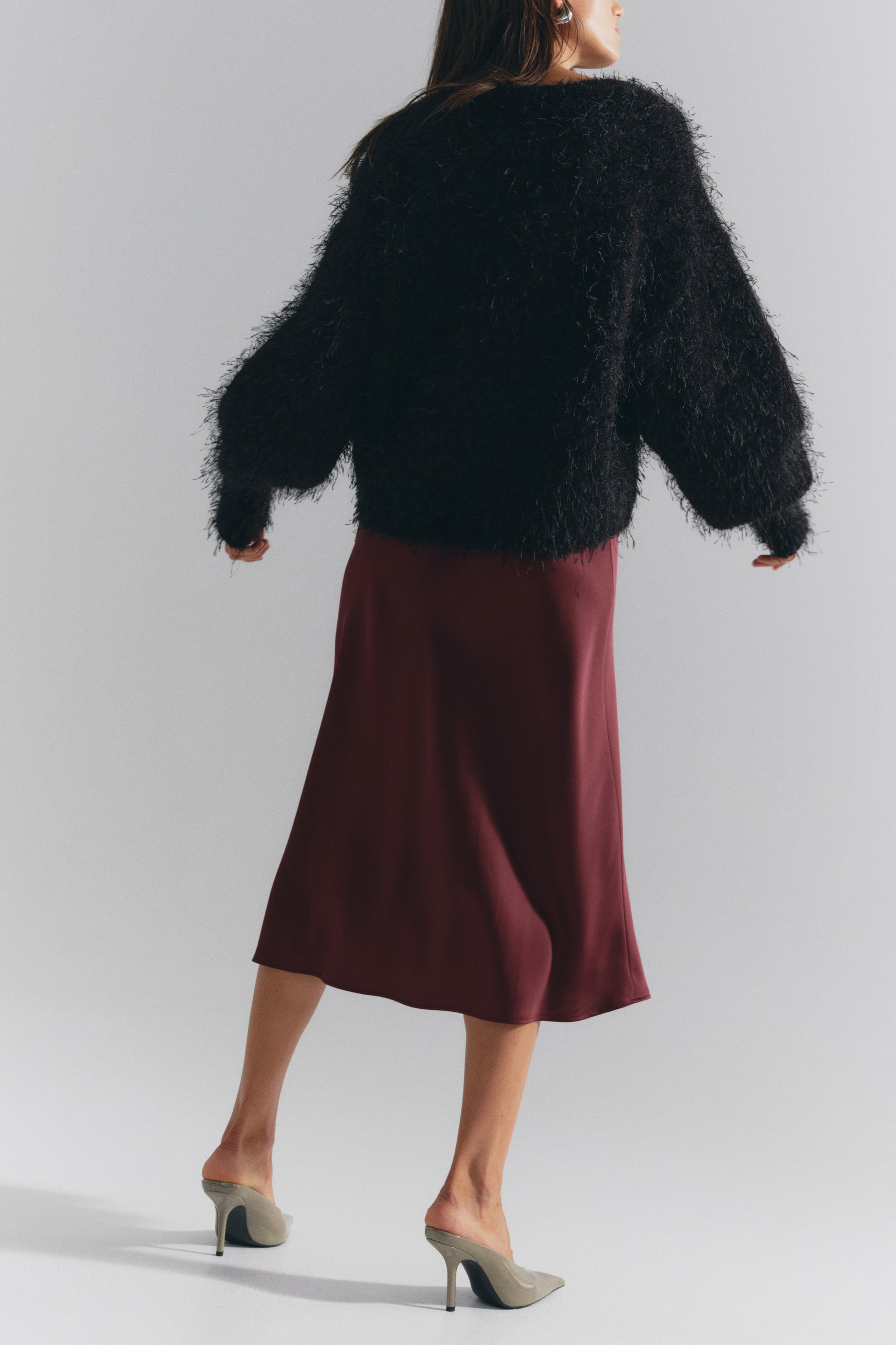 View larger image: A calf-length burgundy satin skirt with a soft, fluid drape is worn by the model, who is standing and facing away from the camera. The skirt is styled with a fluffy, textured black long-sleeved sweater and light green pointed-toe heeled mules.