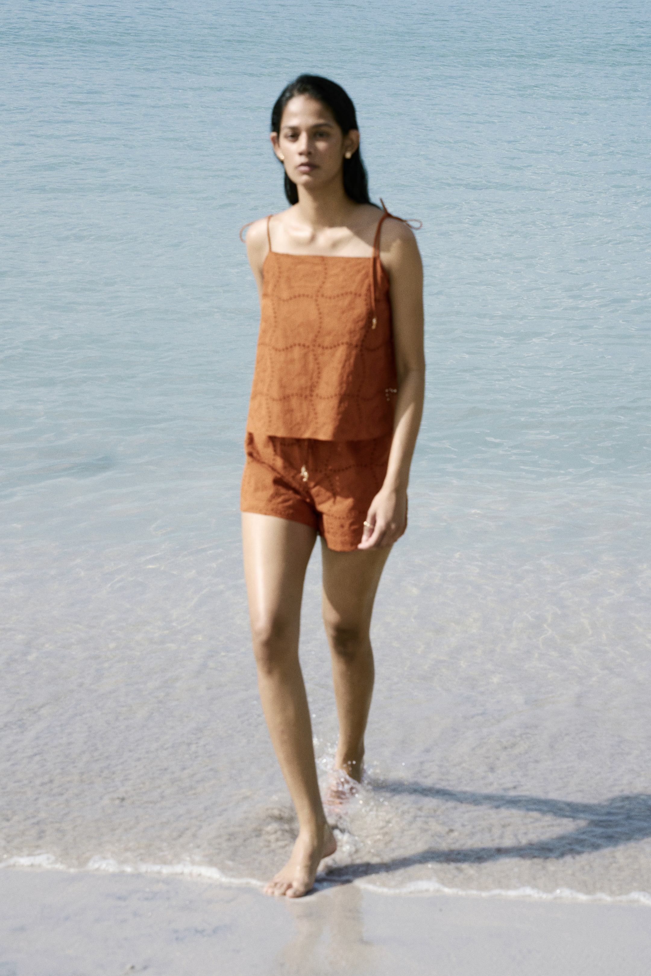 View larger image: Presenting an orange two-piece ensemble, comprising a square-neck camisole and matching shorts, both showcasing a perforated geometric design. The camisole features spaghetti straps, with one strap elegantly tied in a bow, while the shorts include an elasticated drawstring waistband. The outfit is displayed facing forward.
