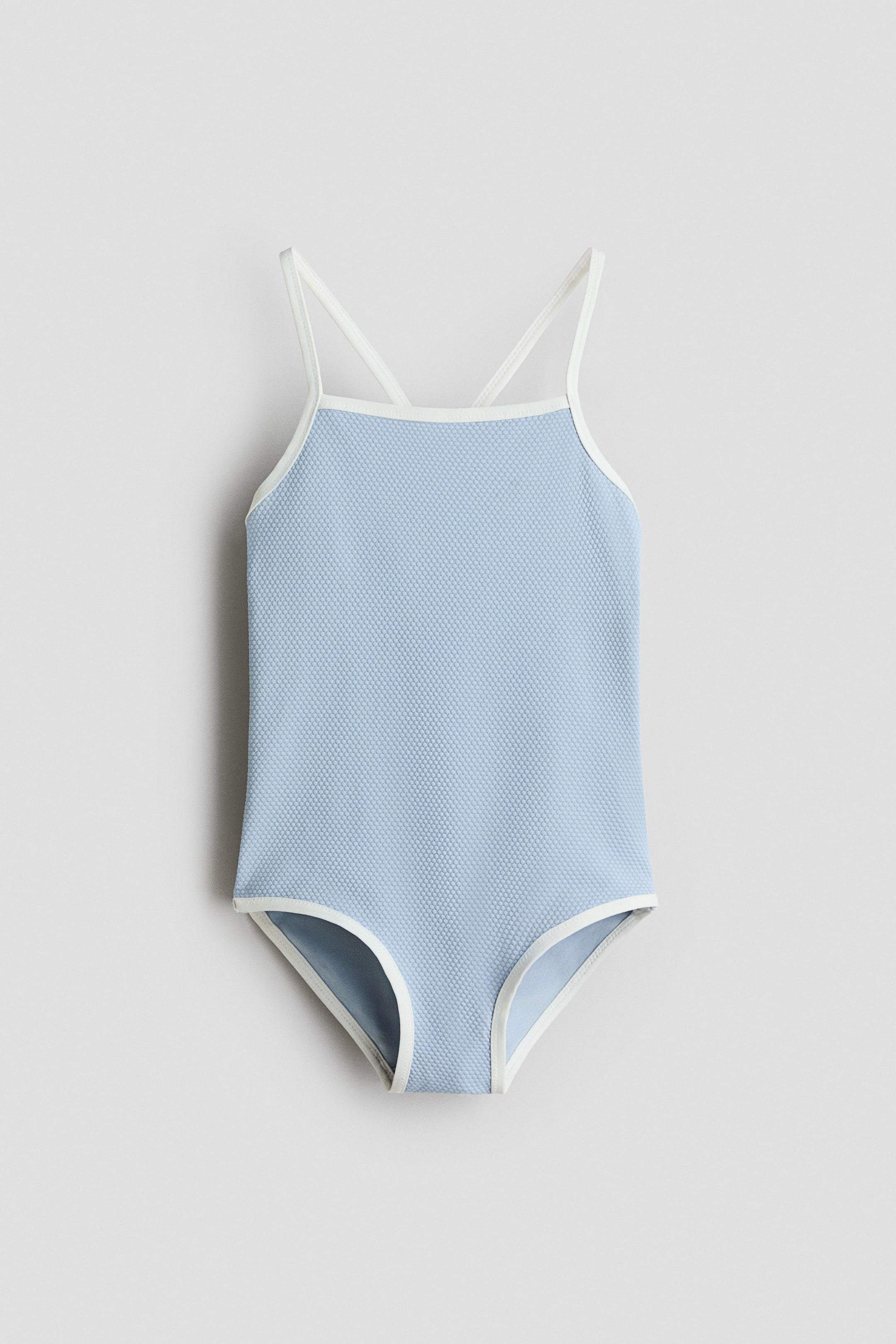 View larger image: A light blue one-piece swimsuit features a textured waffle-knit fabric with white trim outlining the neckline, armholes, and high-cut leg openings. It showcases thin white cross-back straps.