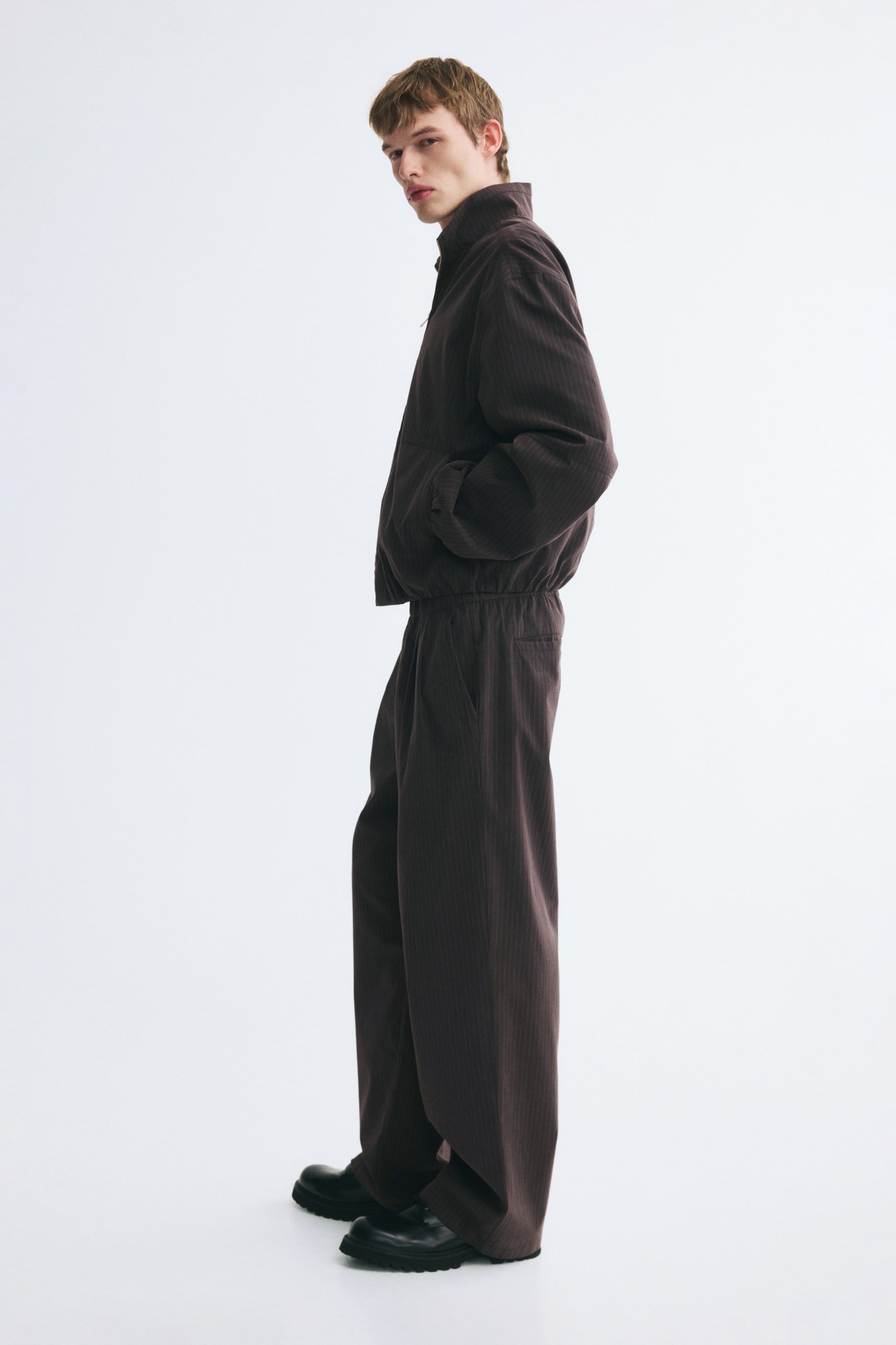 View larger image: Dark brown cotton-blend pants featuring a relaxed fit, straight legs, and subtle vertical texture are being worn by the model. The pants are styled with a matching zip-up jacket and chunky black shoes.