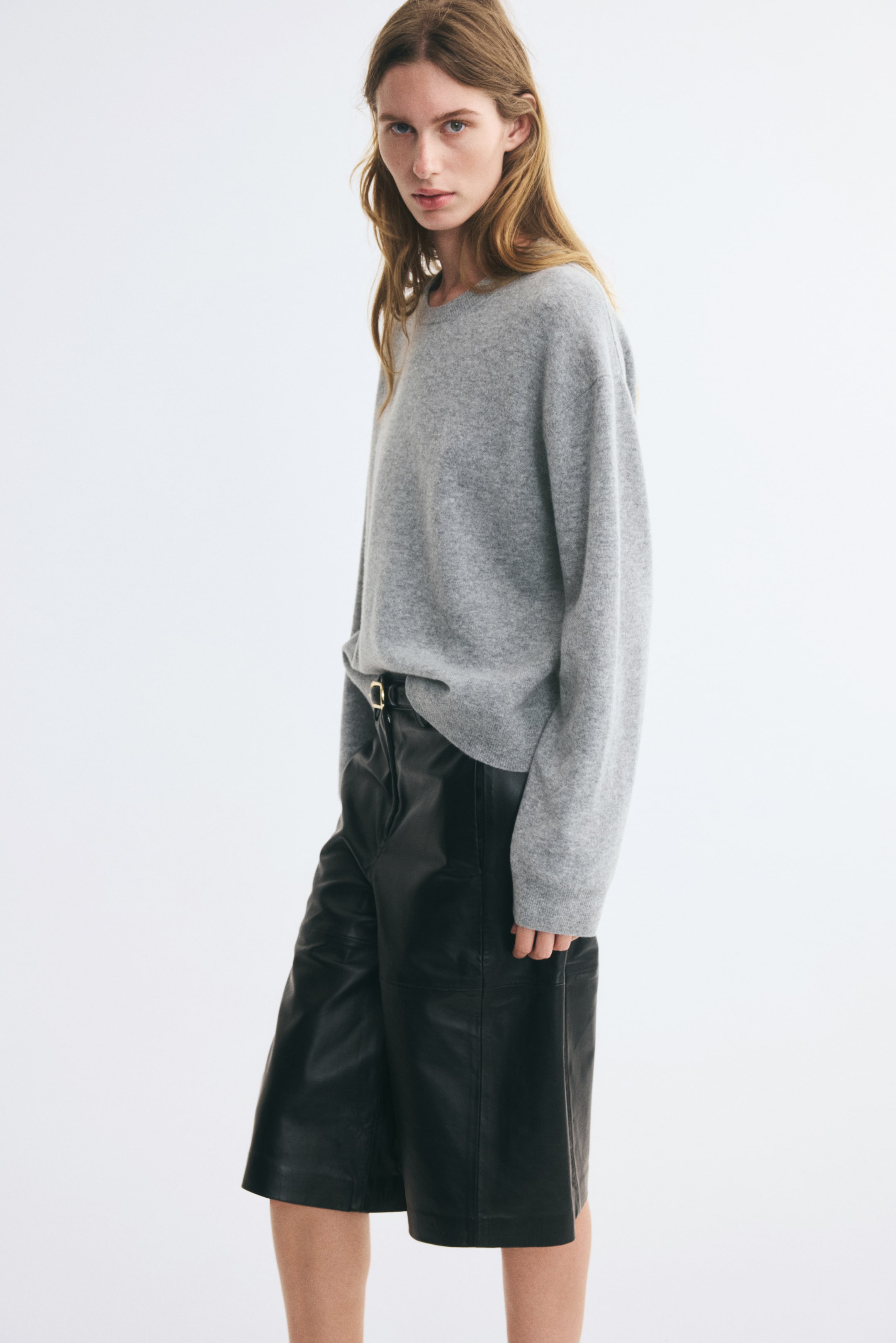 Zobacz większy obraz: Model ma na sobie oversize’owy, szary, dzianinowy sweter wełniany z okrągłym, prążkowanym dekoltem, obniżoną linią ramion i długimi rękawami. Sweter jest częściowo wpuszczony w czarne skórzane szorty.