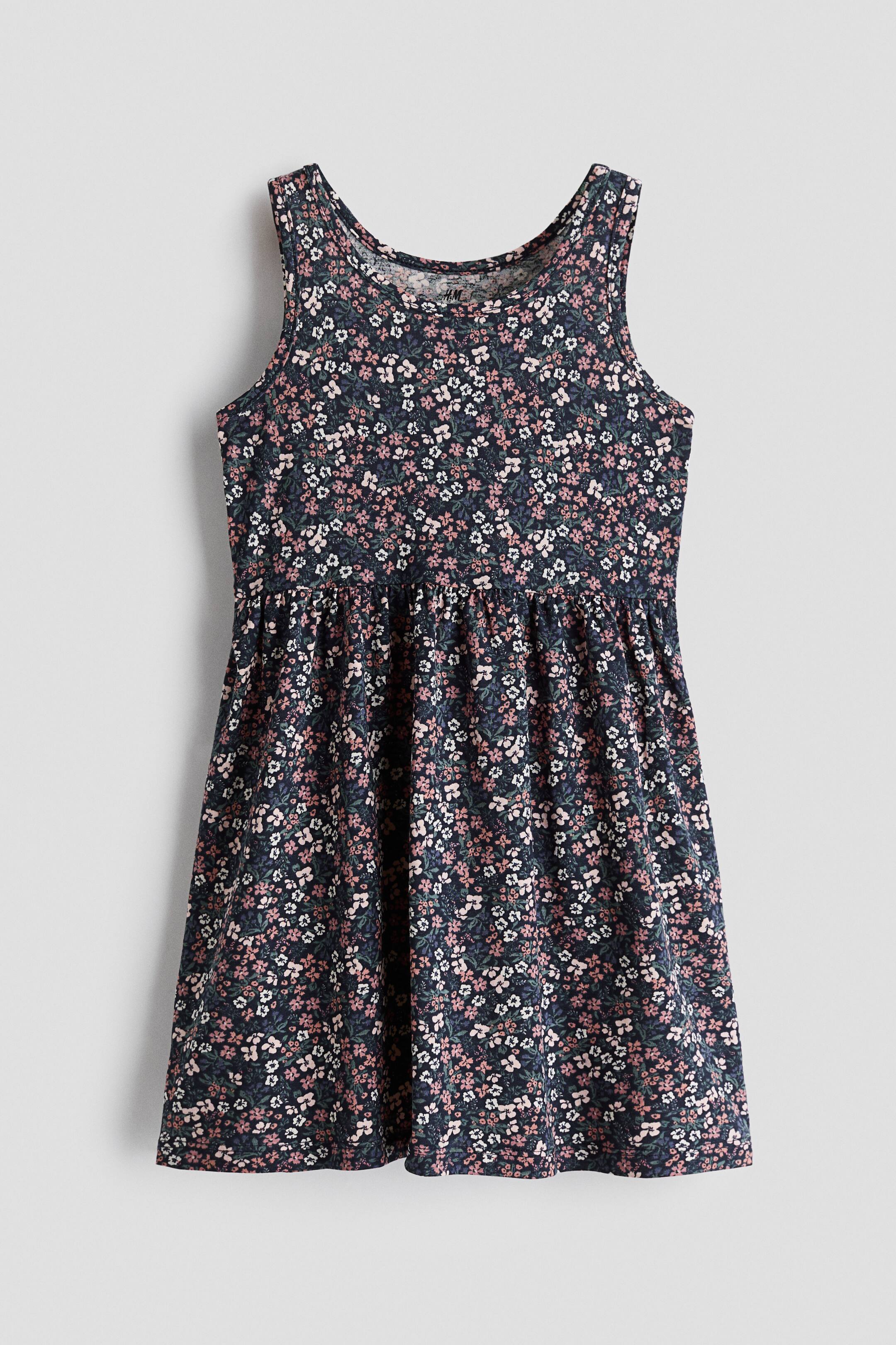 View larger image: A sleeveless jersey dress is shown from the front, featuring an all-over floral print of small pink and white blossoms on a dark blue background. The dress features a round neckline, a gathered seam at the waist, and a flared skirt.