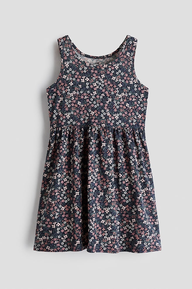 A sleeveless jersey dress is shown from the front, featuring an all-over floral print of small pink and white blossoms on a dark blue background. The dress has a round neckline, a gathered seam at the waist, and a flared skirt. - 1